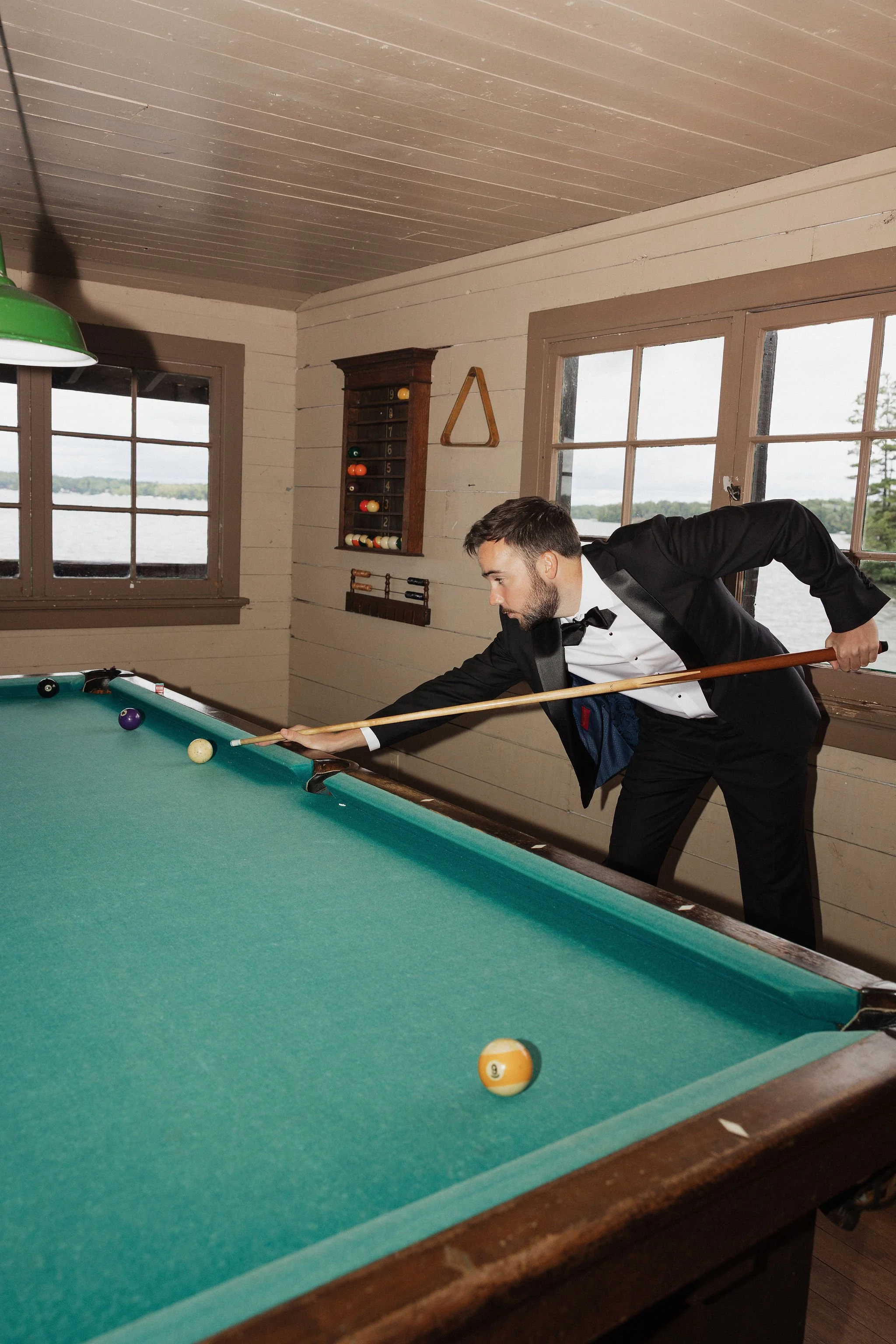 A man in a tuxedo playing billiards inside a rustic wooden room by a lake, aiming to hit the cue ball.