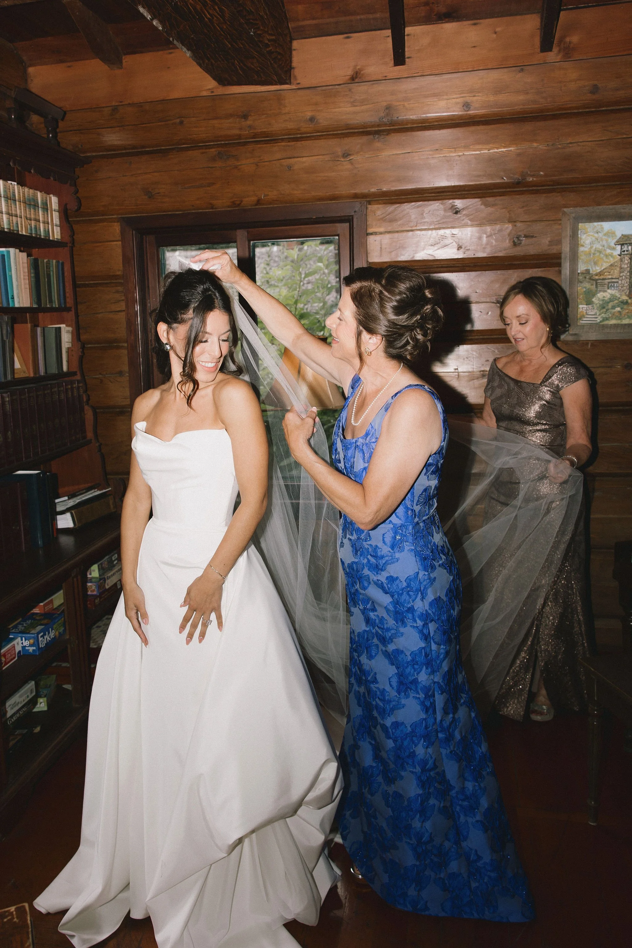 A bride in a white wedding dress is smiling as an older woman in a blue lace dress adjusts her veil in a wooden room. Another woman in a sparkly dress helps hold the veil.