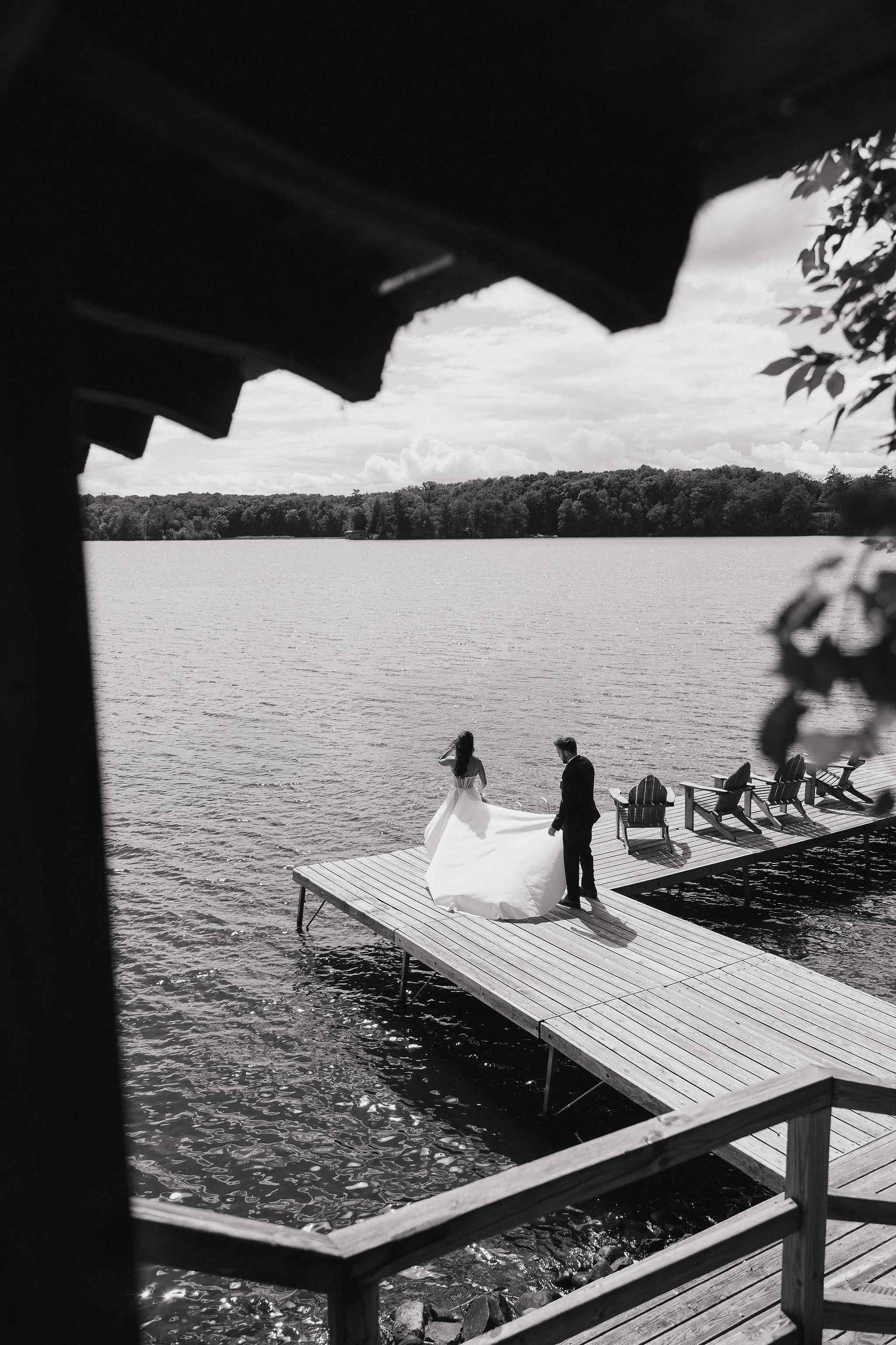 A black and white photo of a bride and groom standing on a dock by a lake, with the bride in a wedding dress and the groom in a suit, surrounded by empty deck chairs.