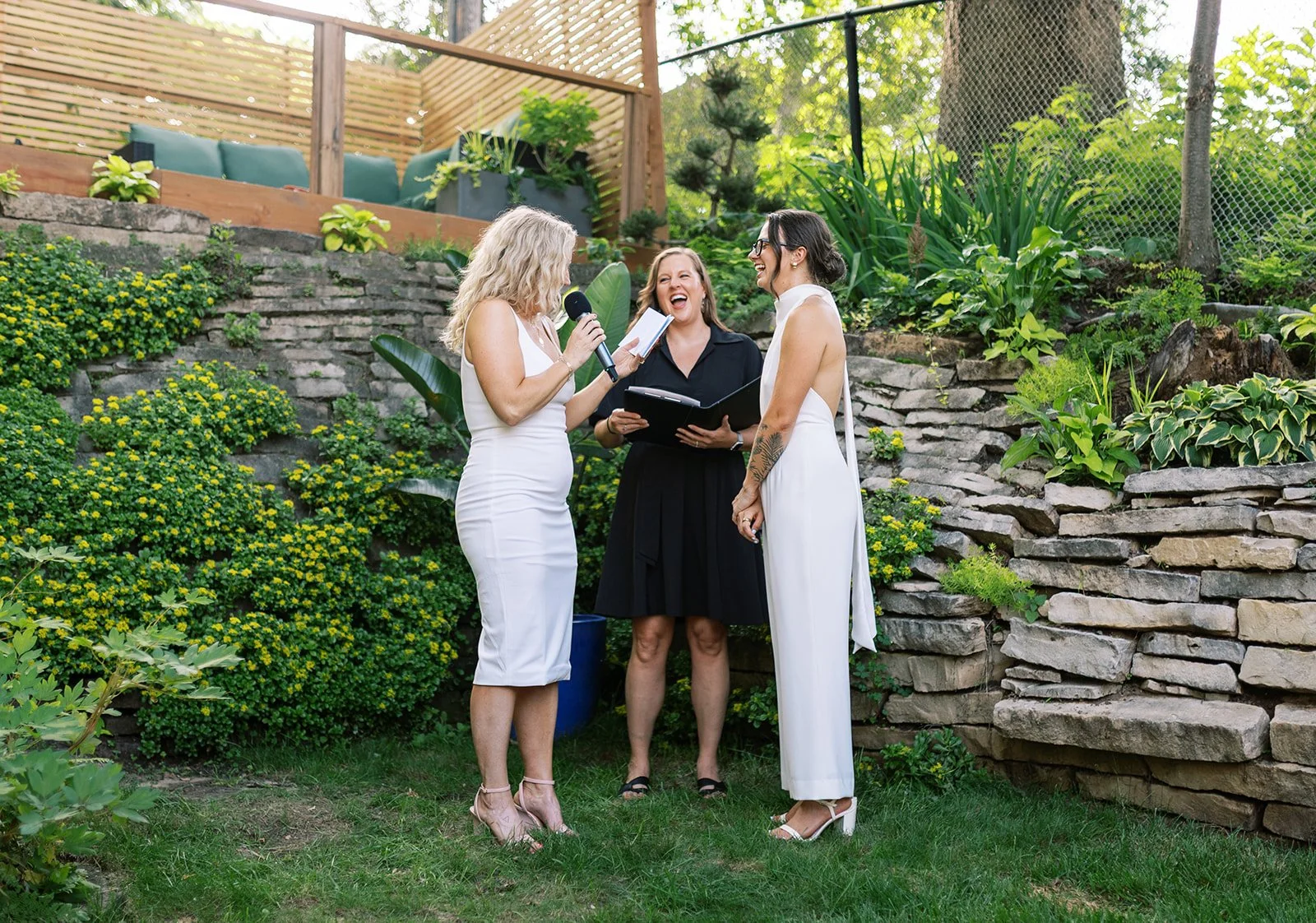 A woman in a white dress is getting married outdoors, standing with a woman in a black dress and a woman in a white jumpsuit. The officiant is reading the vows while everyone laughs, surrounded by green plants and stone steps.