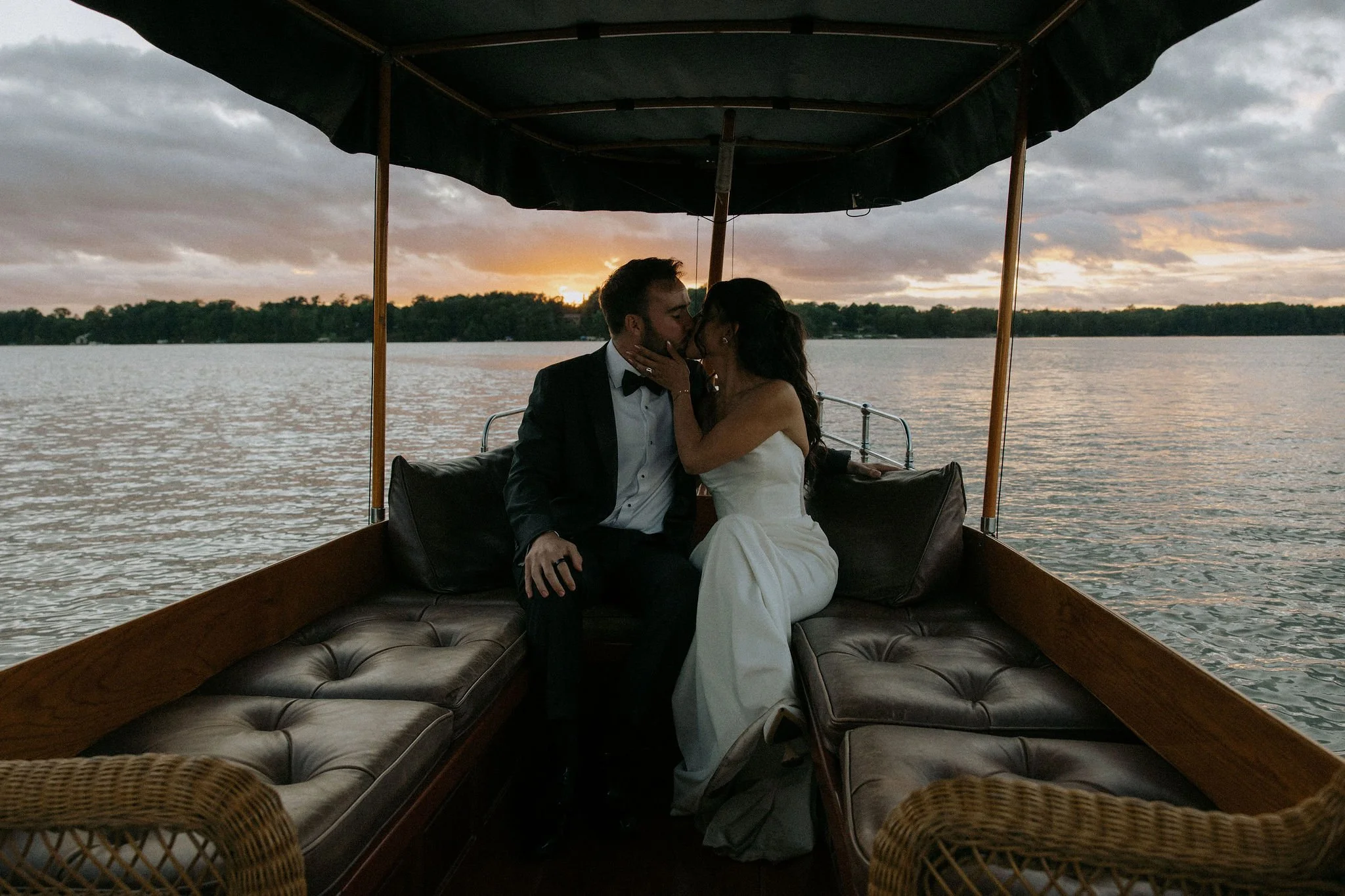 A man in a tuxedo and a woman in a white dress sharing a kiss on a boat at sunset over a body of water with a tree-lined shore in the background.