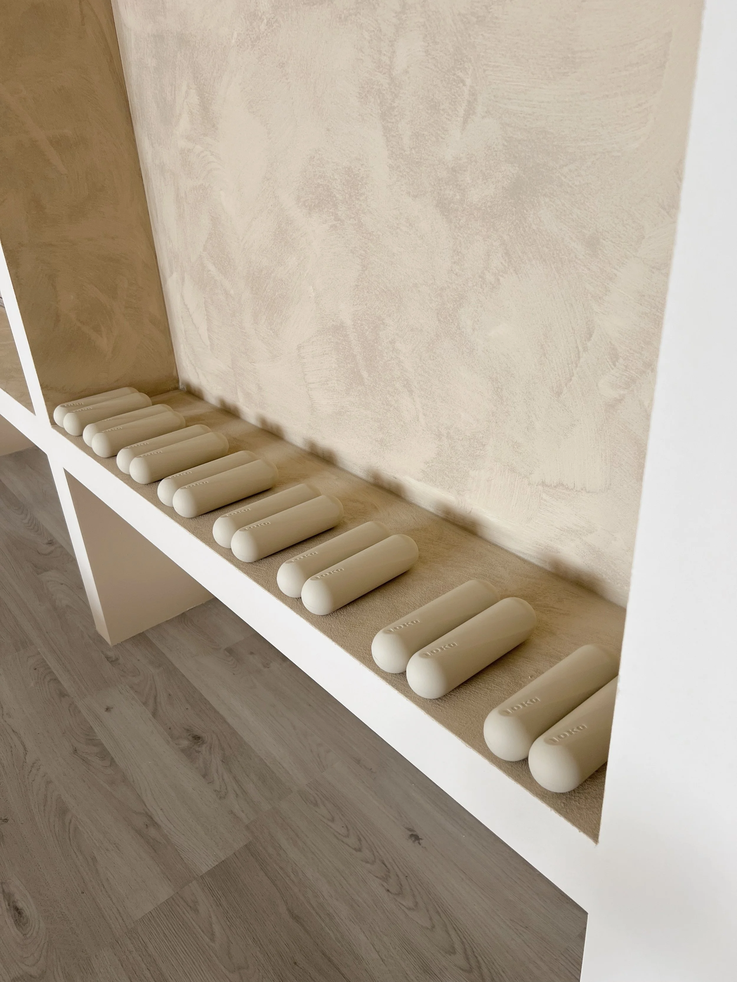 Una mensola bianca con numerosi cilindri di sapone disposti in fila, sul pavimento in legno e contro una parete beige.