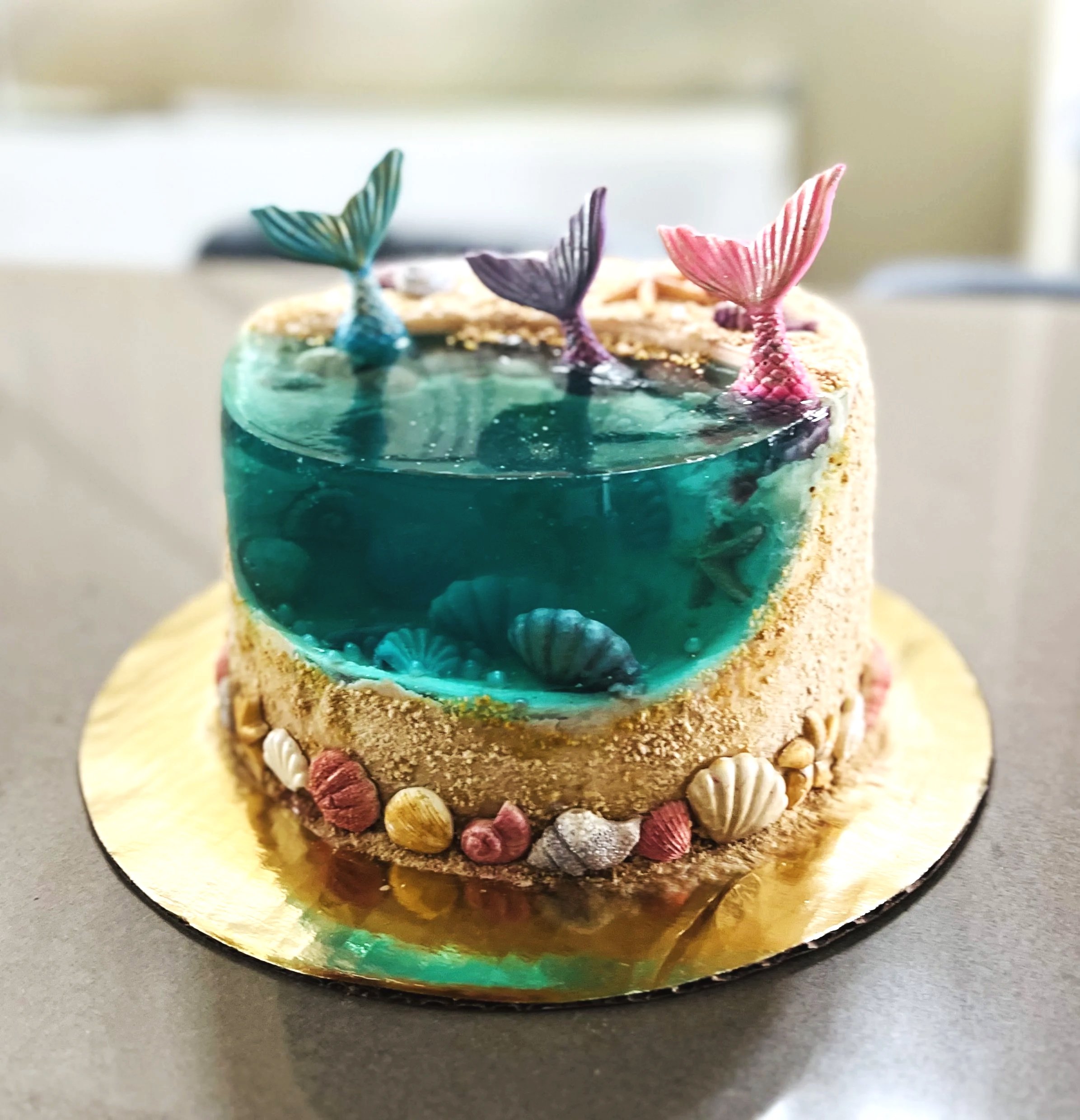 A cake designed to look like an underwater scene with mermaid tails and seashells, featuring a half-globe shape with blue-green water and marine elements.