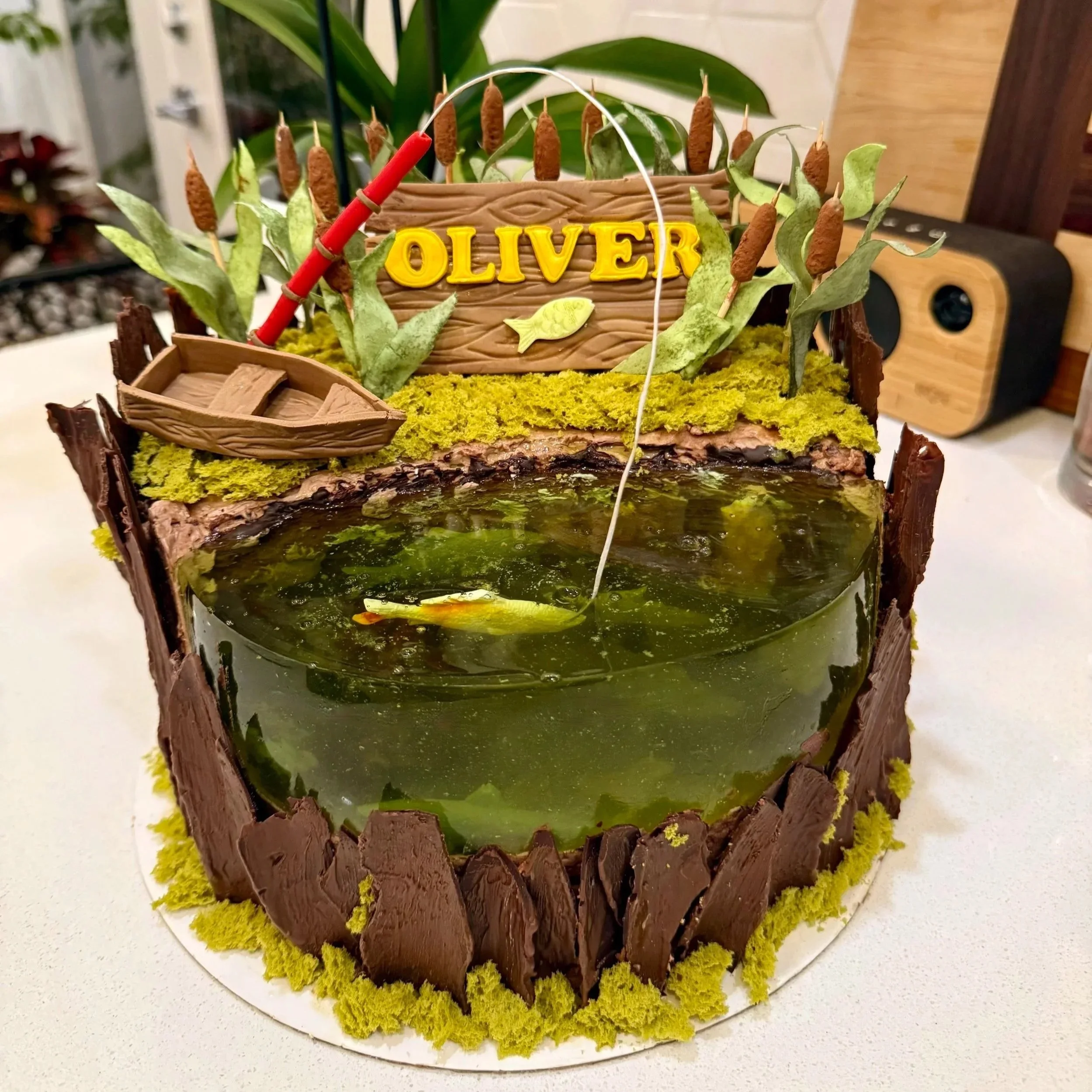 A boat-shaped cake with a pond on top, decorated with moss, chocolate, and fishing-themed accents.