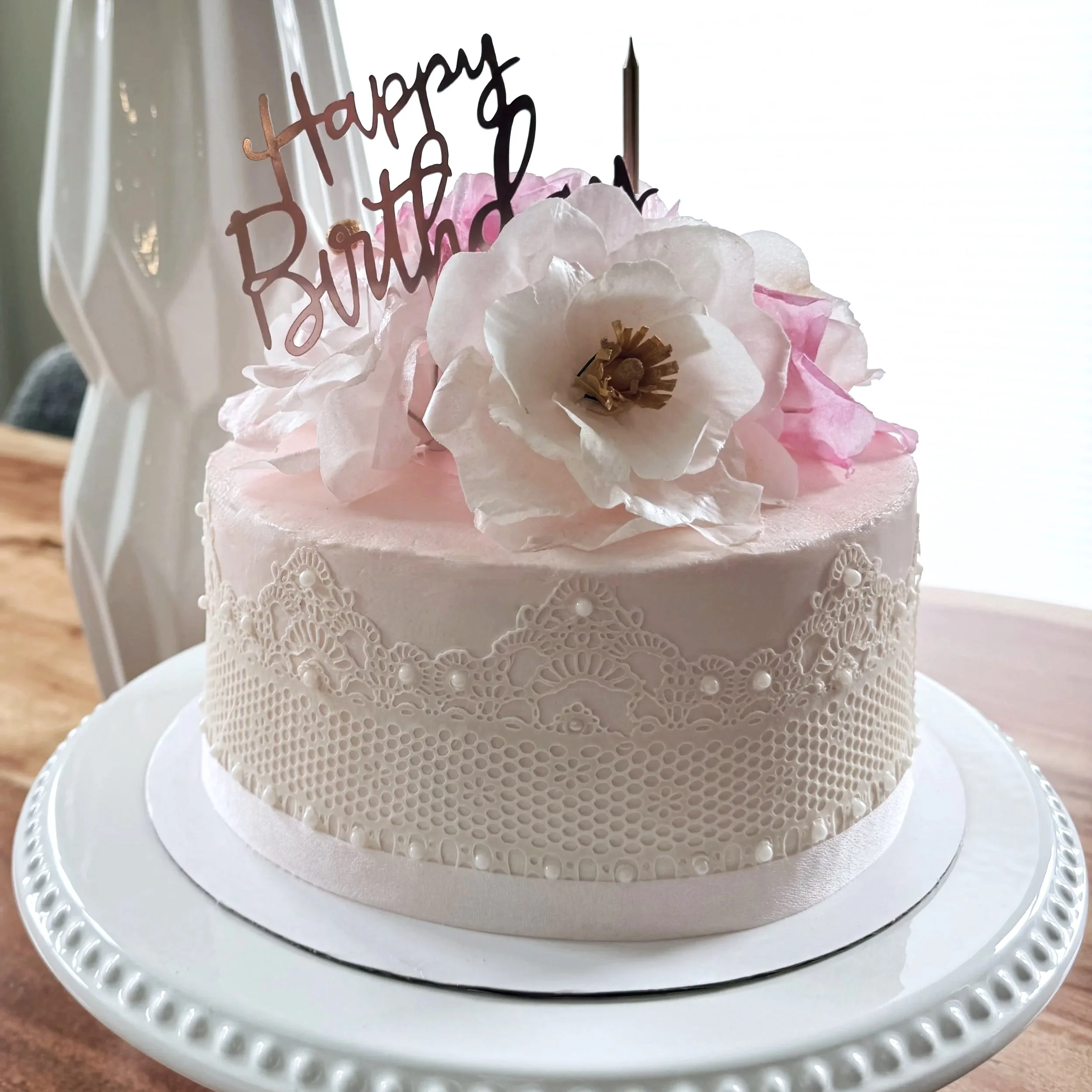 Pink and white birthday cake decorated with large flowers and a 'Happy Birthday' topper, placed on a white cake stand.