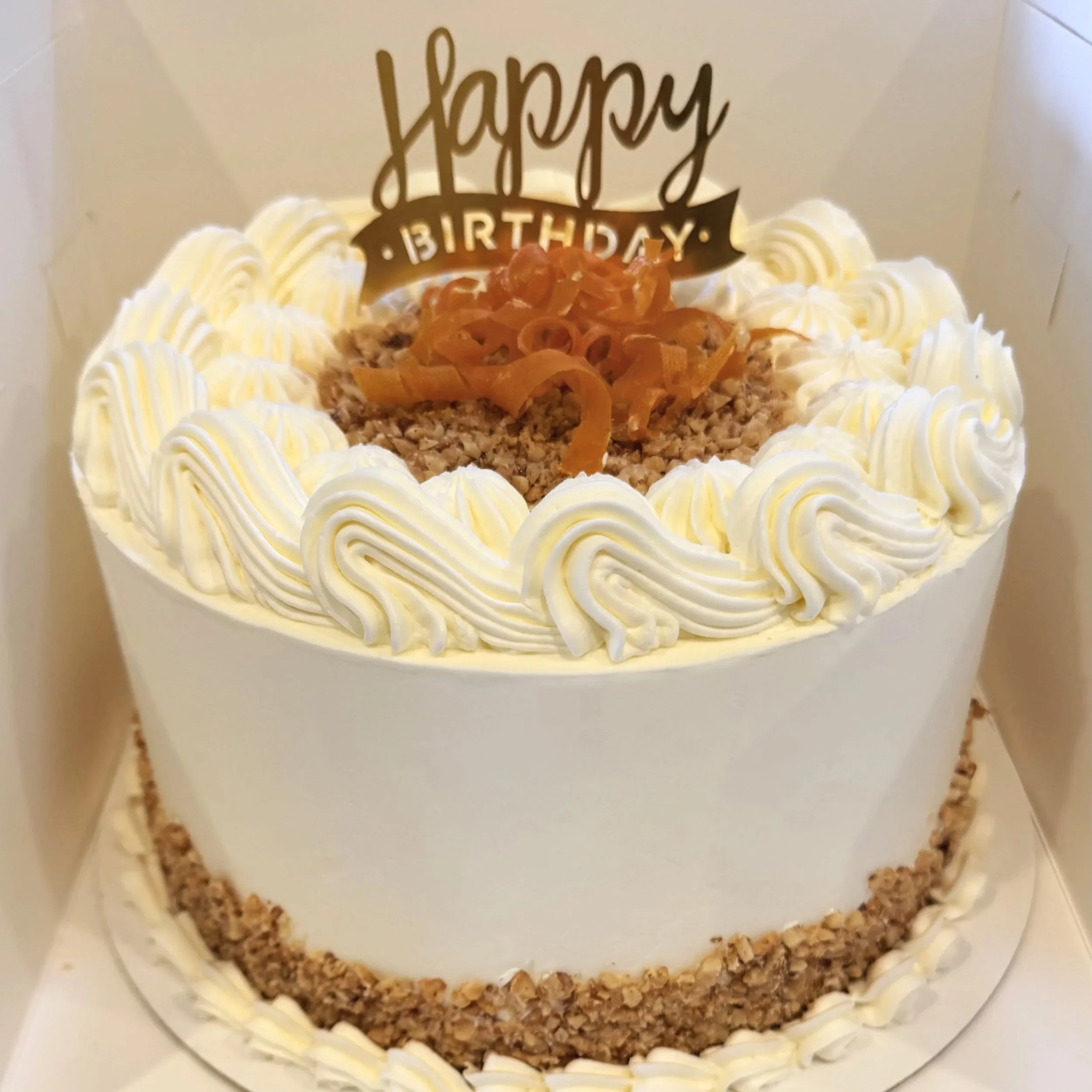 A round birthday cake with white frosting and decorative piping, topped with a "Happy Birthday" sign and caramelized topping, inside a white box.