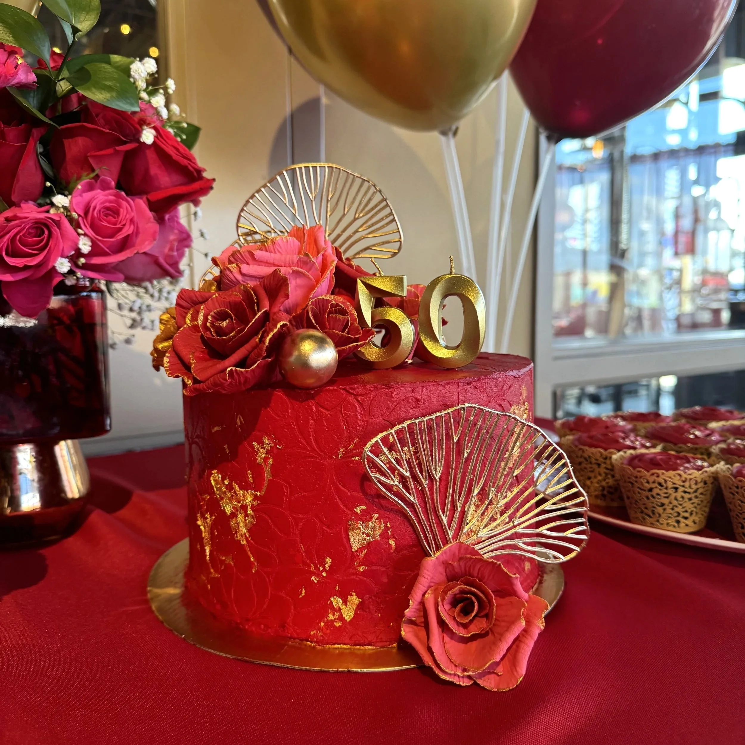 A red cake decorated with golden accents, pink and red flowers, the number 50 in gold, and gold fan-shaped embellishments, set on a red tablecloth