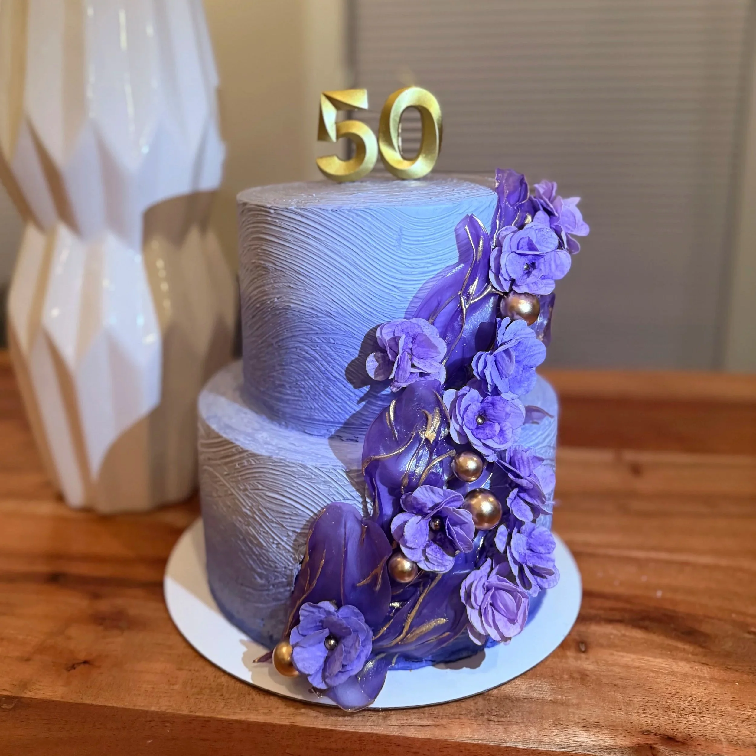 A two-tiered cake with a textured silver-gray icing, decorated with purple flowers and gold accents. A gold '50' topper is placed on top.