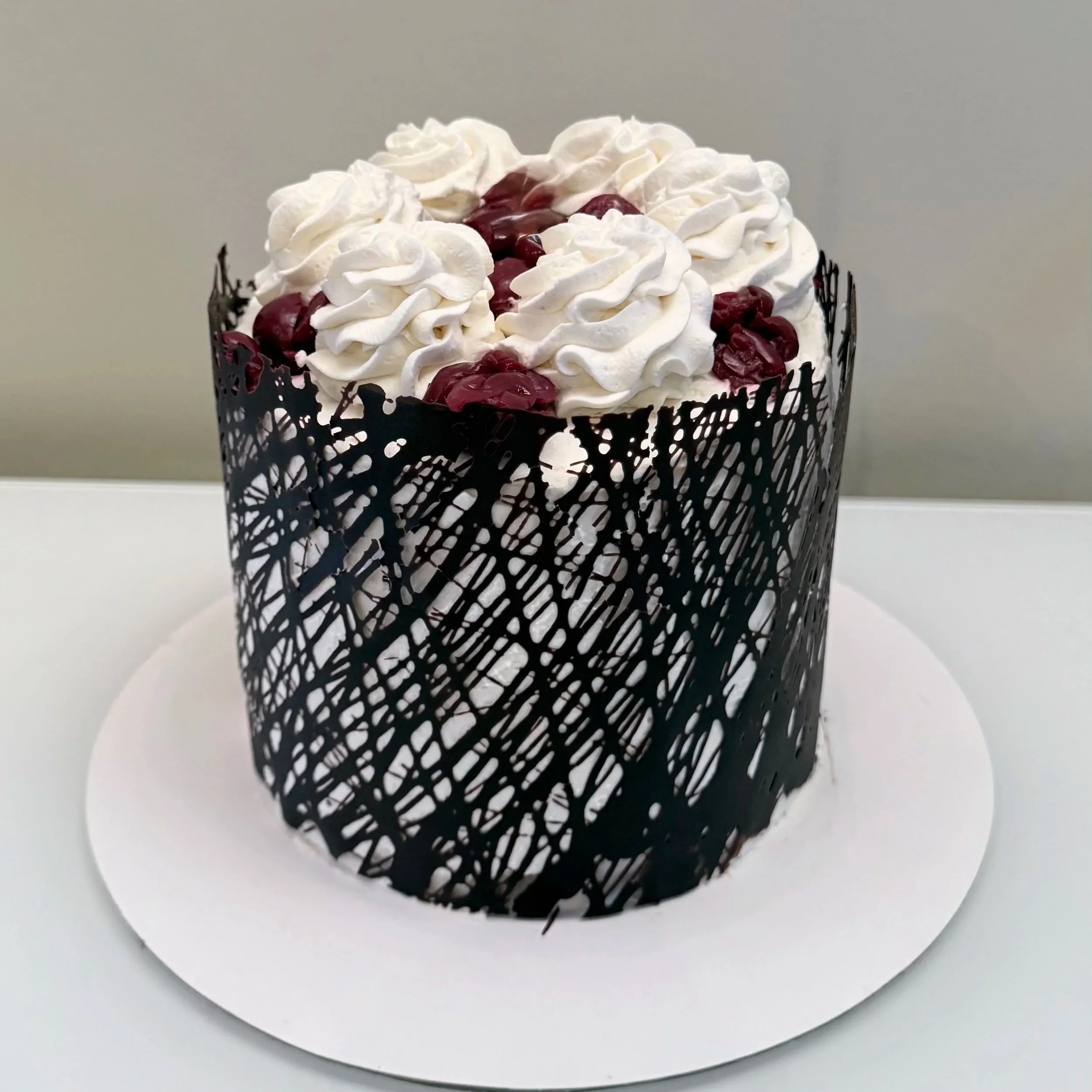 Chocolate cake decorated with white whipped cream and cherry topping, encased in a black decorative lattice cake protector, on a white cake board.