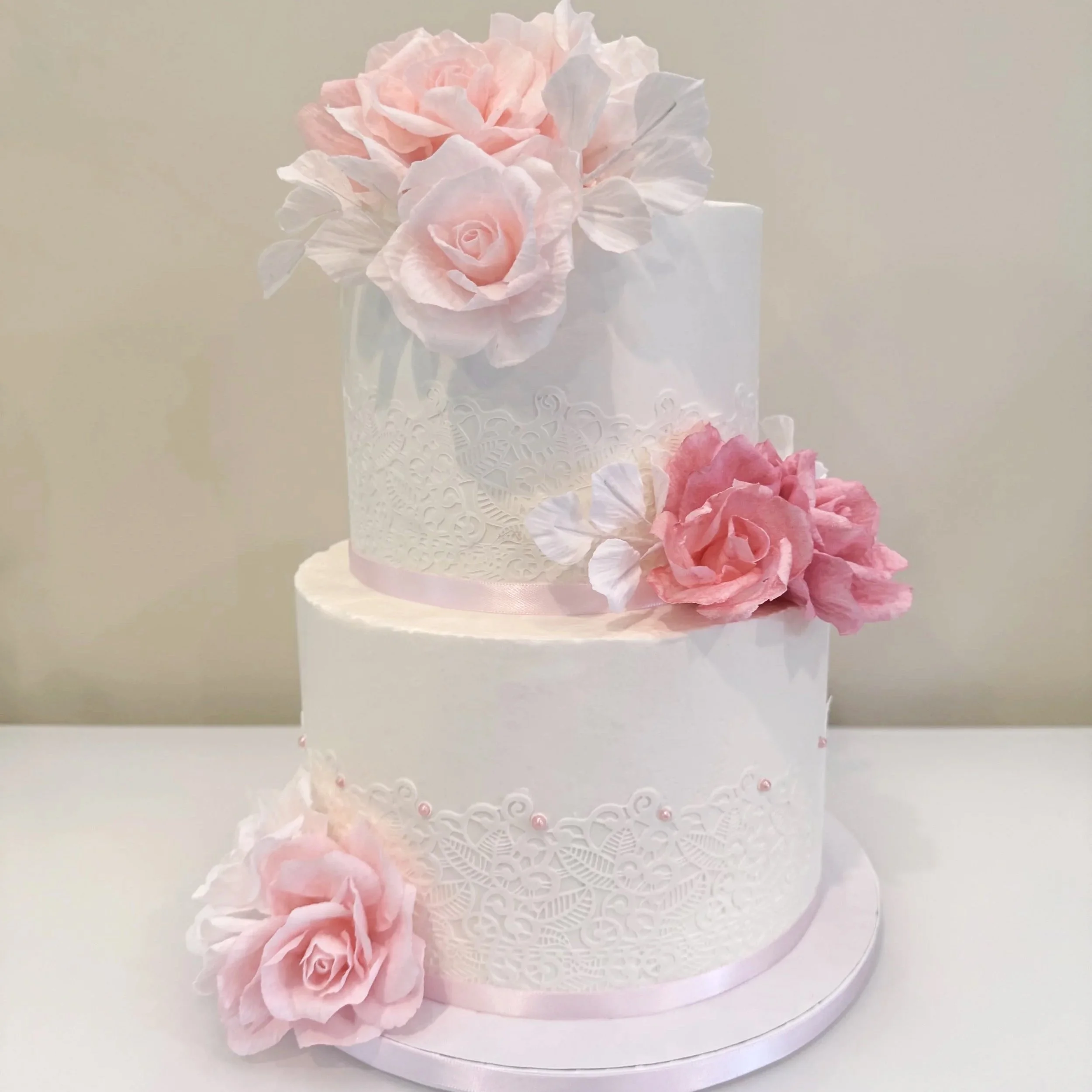 A two-tier white wedding cake decorated with pink roses and intricate lace patterns, with additional roses on the base and top tier.
