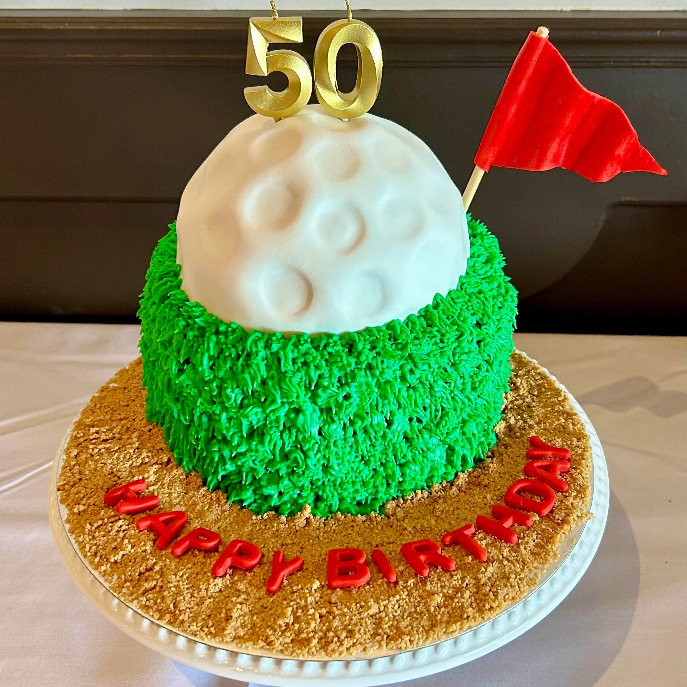 A birthday cake designed to look like the moon, with a white spherical top and crater-like indentations, decorated with green piped frosting around the base, a red flag, and a gold '50' topper, with 'HAPPY BIRTHDAY' written in red letters on a crumb-
