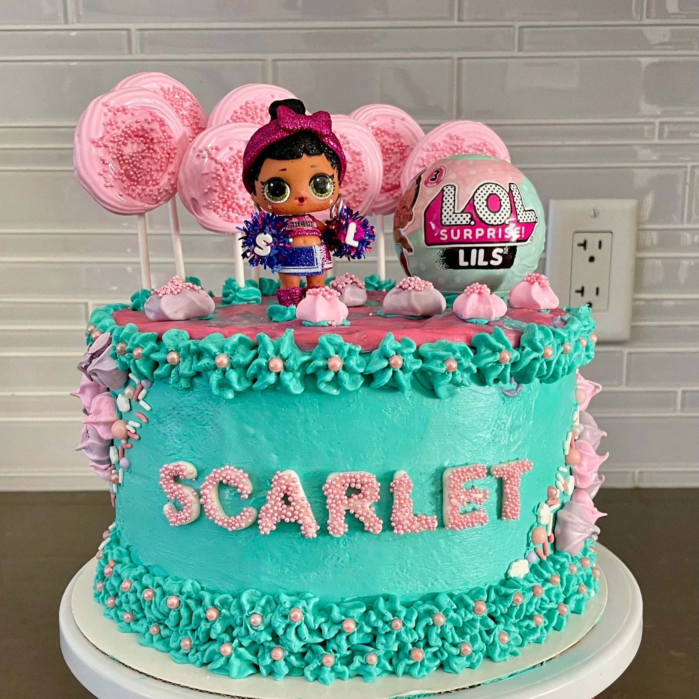 A colorful birthday cake with turquoise frosting, pink and purple decorations, a doll figure, pink lollipops, and a ball with LOL Surprise logo, with the name 'SCARLET' written on the front.