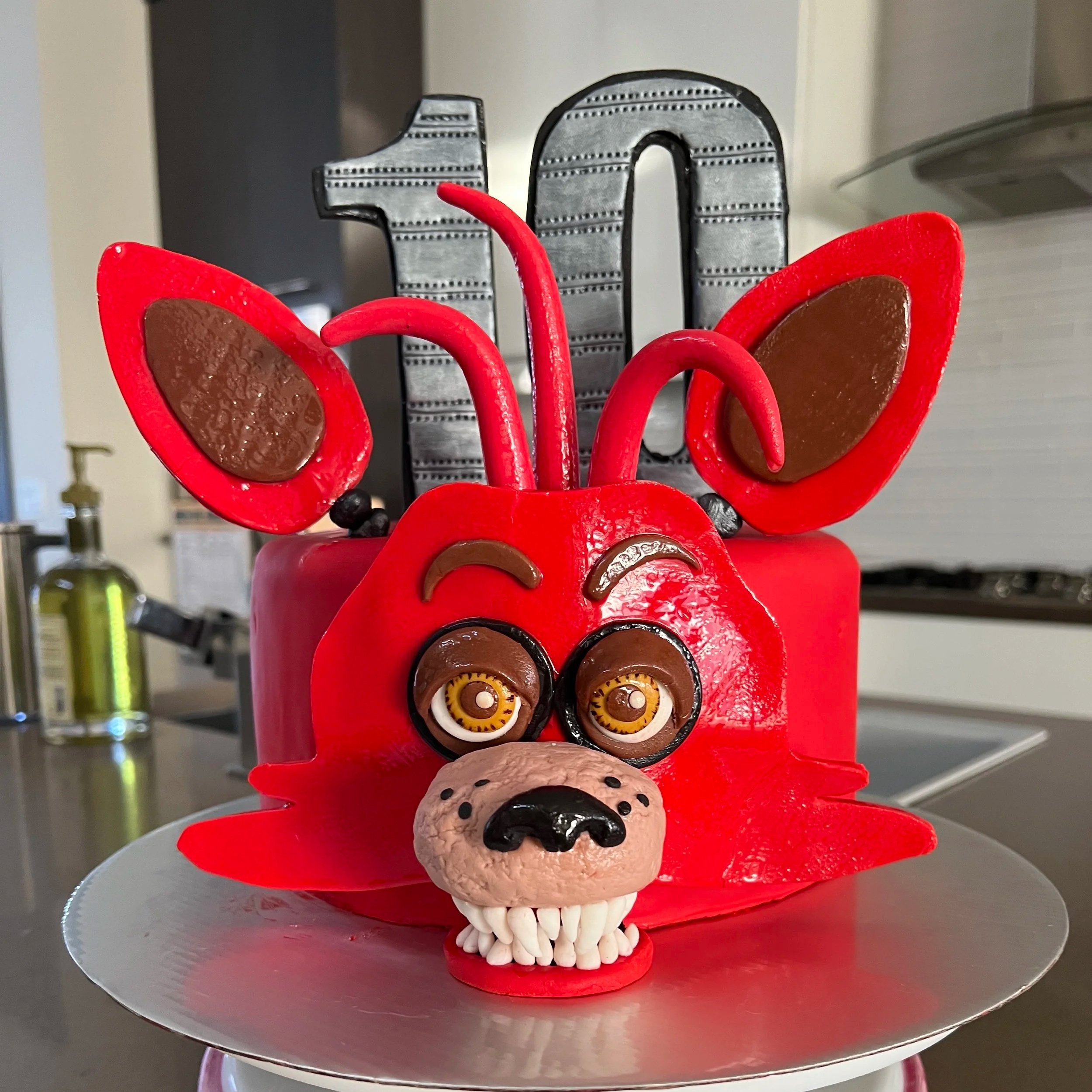 A cake designed to look like a red cartoon dog with large ears, eyes, a nose, a snout with teeth, and a black "10" decoration on top, made for a 10th birthday celebration.