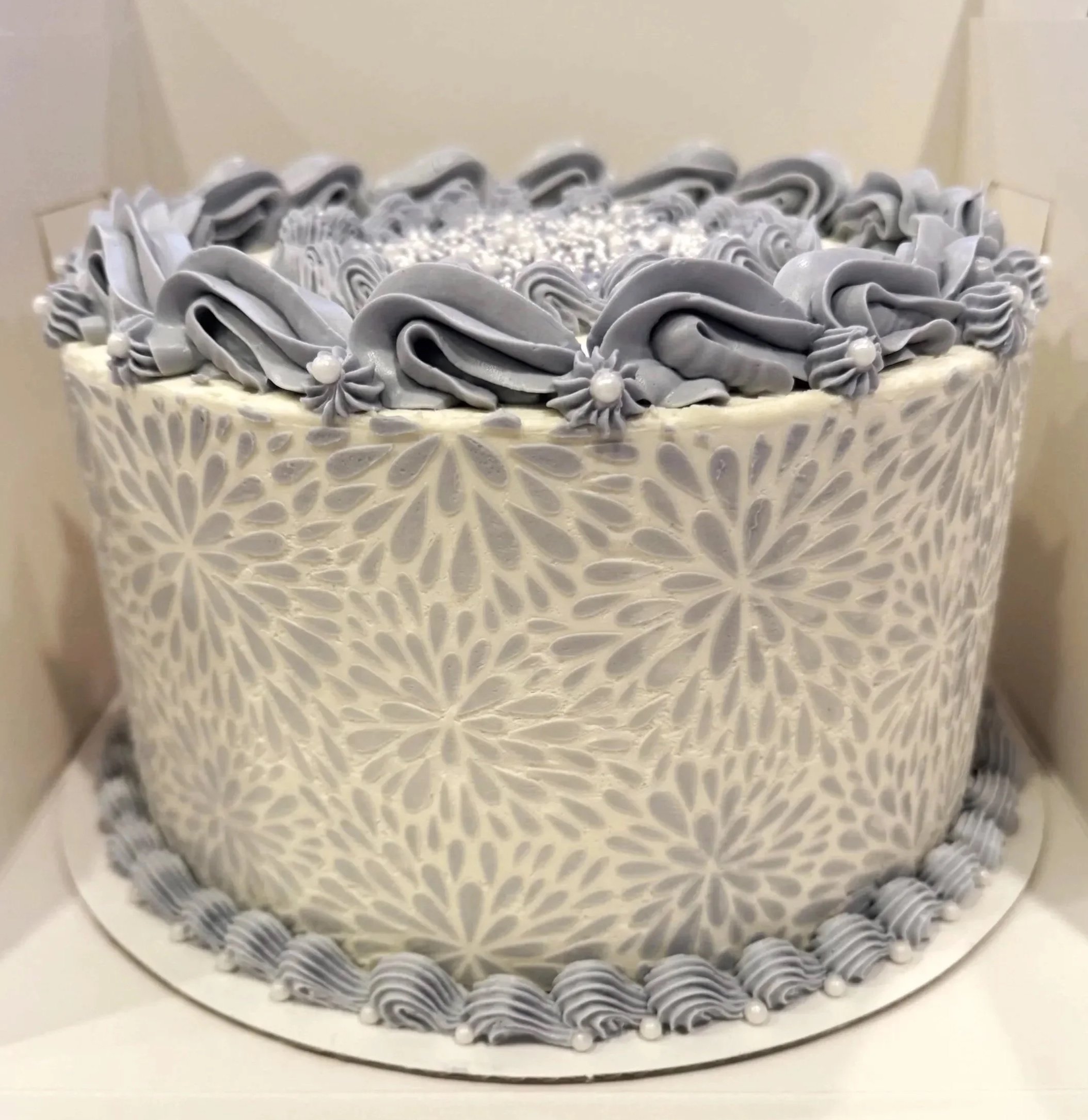 A round wedding cake decorated with white and gray frosting, floral patterns on the sides, and gray frosting swirls with pearl accents on top.