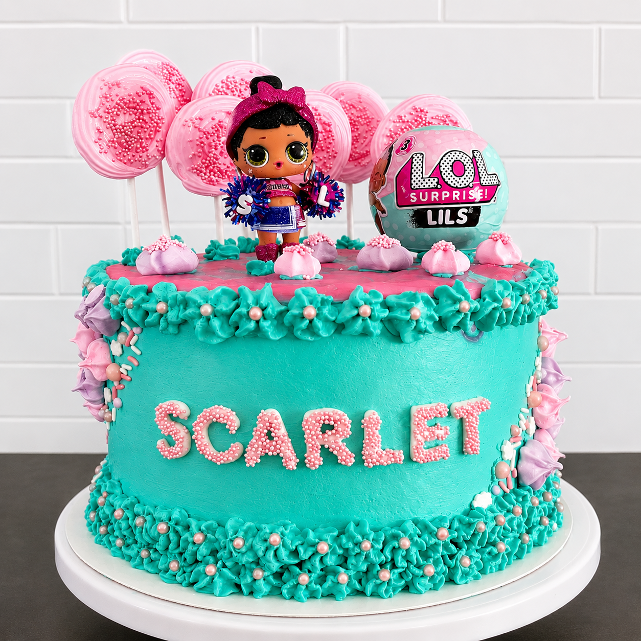 A bright and playful custom birthday cake featuring vibrant turquoise buttercream, pink candy accents, and an adorable LOL Surprise theme. Decorated with piped ruffles, cake pops, sprinkles, and personalized details, this fun design is topped with a 