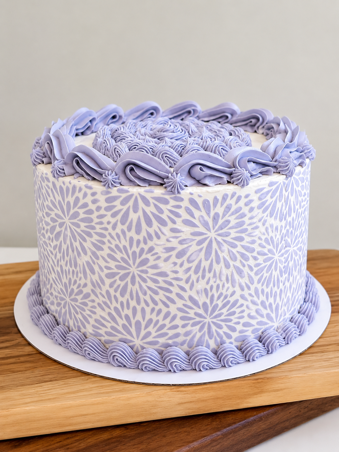 An elegant buttercream cake featuring a soft lavender and white floral stencil design with delicate piped borders and textured top detailing. Finished with vintage-inspired piping and a clean modern palette, this custom cake is perfect for birthdays,
