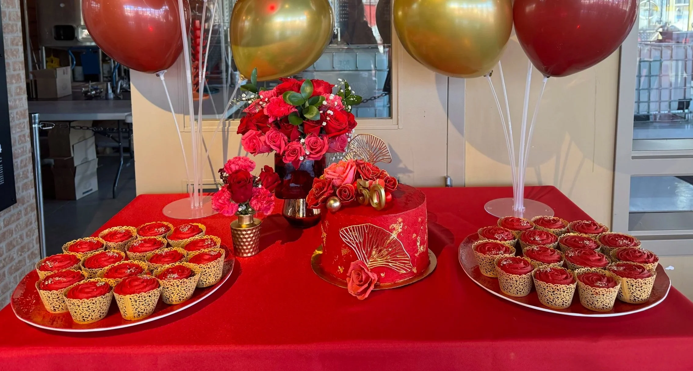 Decorative table setup with a red birthday cake, gold and red balloons, and floral arrangements, celebrating a 50th birthday.