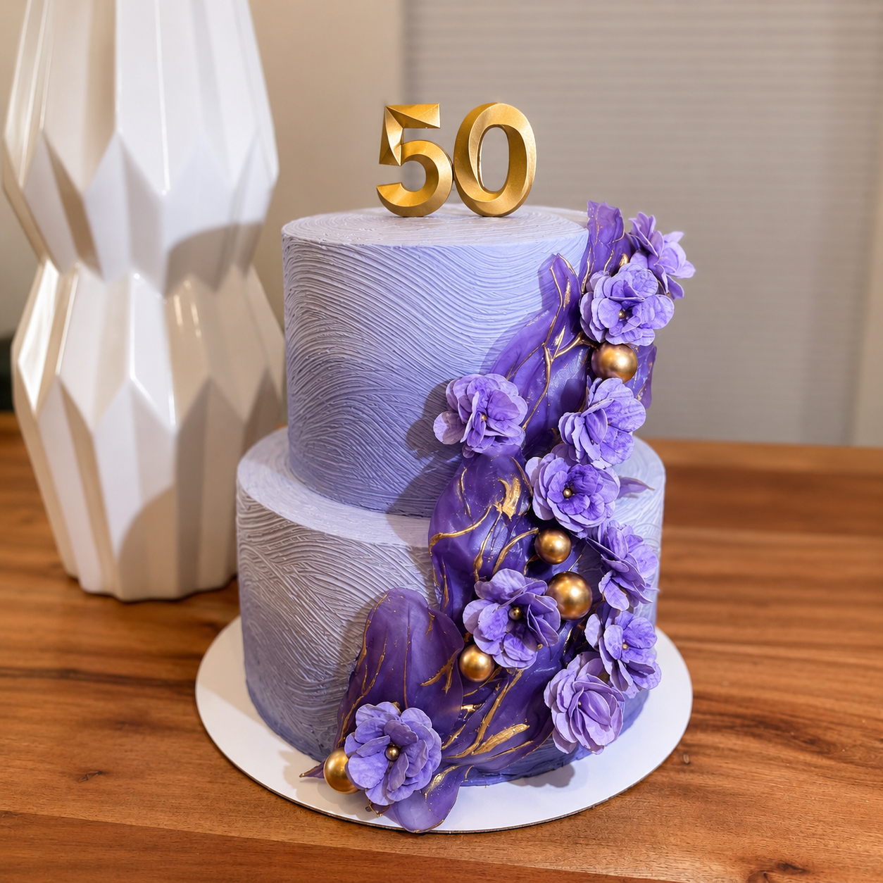 A sophisticated two-tier buttercream cake featuring textured lavender finishes, dramatic rice paper sails, and luxurious gold accents. Cascading handcrafted florals and metallic gold details create a bold yet elegant design, finished with a modern go