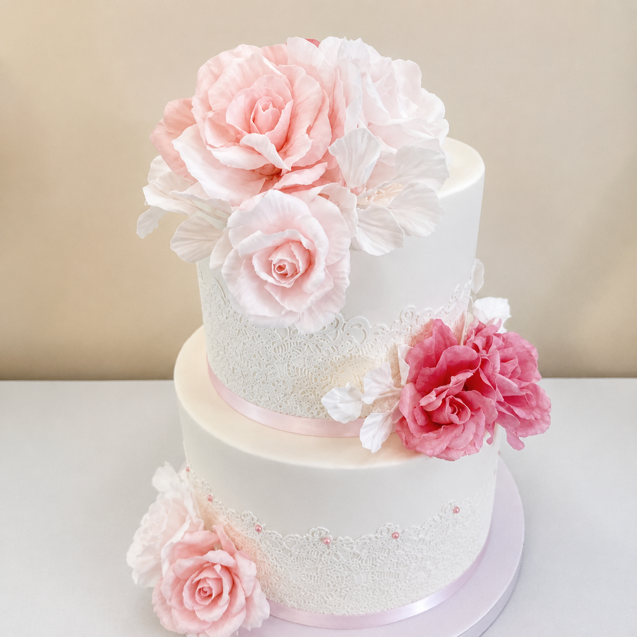 Two-tier white wedding cake decorated with pink and white sugar flowers, topped with large pink roses.