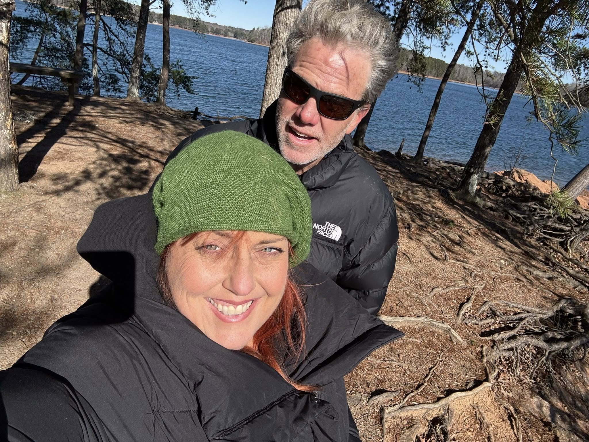 A smiling woman wearing a green beanie and black jacket takes a selfie in a wooded outdoor area near a lake, with a man in sunglasses and a black jacket behind her, during daytime.