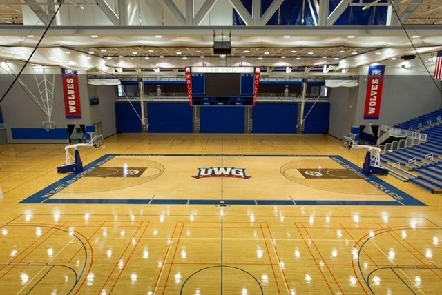 Empty indoor basketball court with wooden floor, UWM logo at center, blue padded walls, and banners that read "WOLVES" hanging from the ceiling.