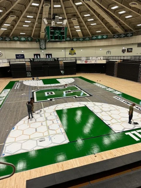 Basketball court in a gymnasium with a green and white logo at the center, surrounded by green and white borders, and two people working on the court.