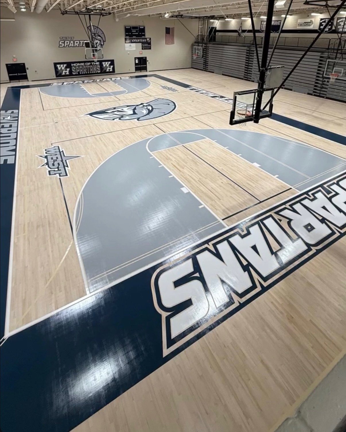 An indoor basketball court with a light wooden floor, blue and white markings, and team logos. The court has a large logo at the center and the words 'GARDENINGS' along the sideline. Bleachers are visible on the right side of the court, and basketbal