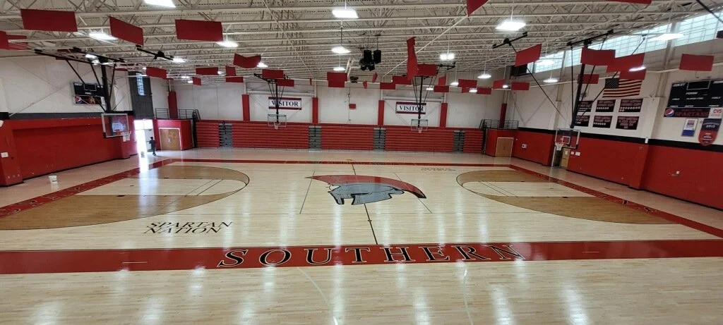 Empty basketball gymnasium with a wooden floor, red walls, and banners hanging from the ceiling, featuring the South Carolina Gamecocks logo and colors.