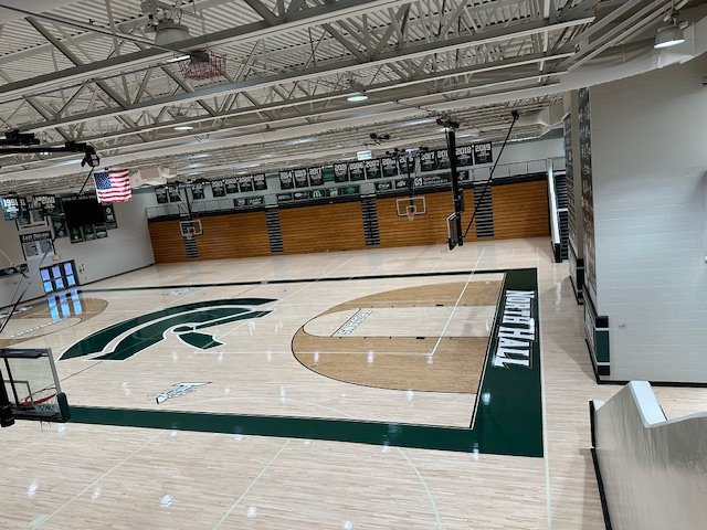 Empty indoor basketball court with Michigan State Spartan logo at the center, surrounded by wood paneling, basketball hoops, and banners, with an American flag hanging from the ceiling.