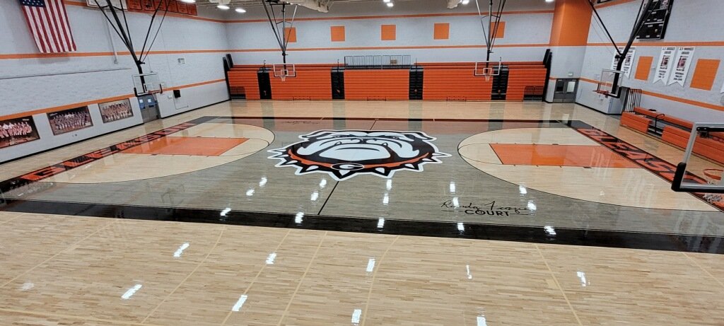 An empty basketball court featuring a bulldog mascot logo at the center, with orange and black accents, and bleachers along the walls.