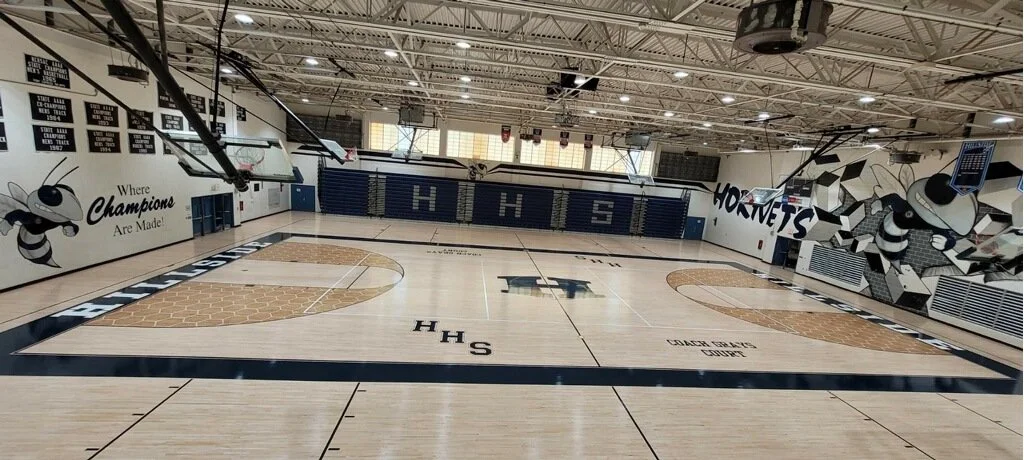 Empty high school gymnasium with basketball court, blue bleachers, and mural art of bees on the walls.