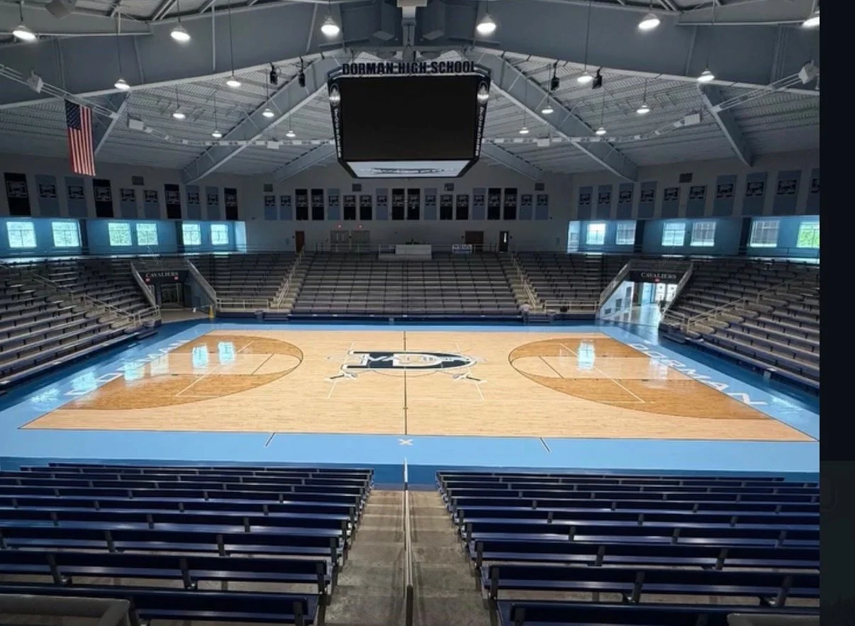 Empty basketball court inside an indoor sports arena with seating on all sides, a hanging scoreboard, and an American flag.