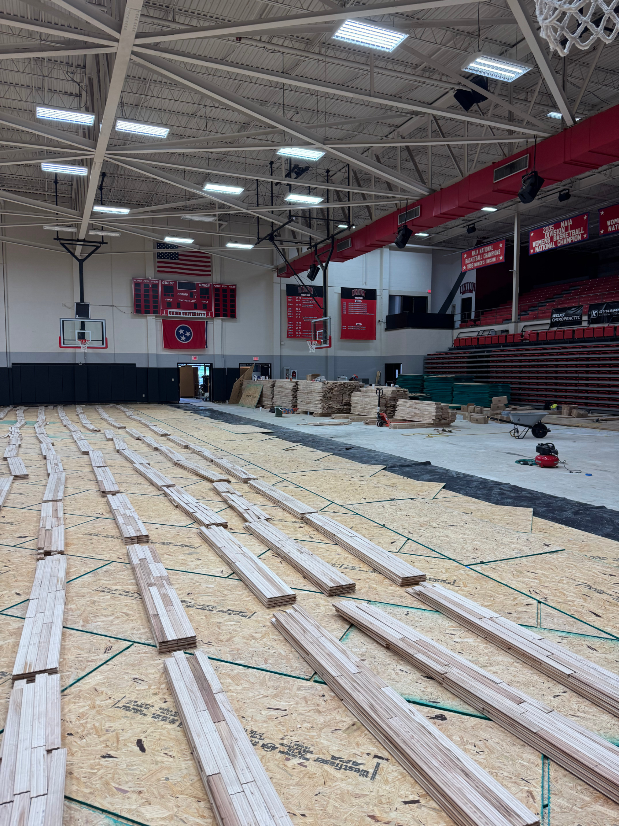 Interior of a gymnasium under renovation, with wooden planks and construction tools on the floor, basketball hoops on the walls, and red banners hanging from the ceiling.