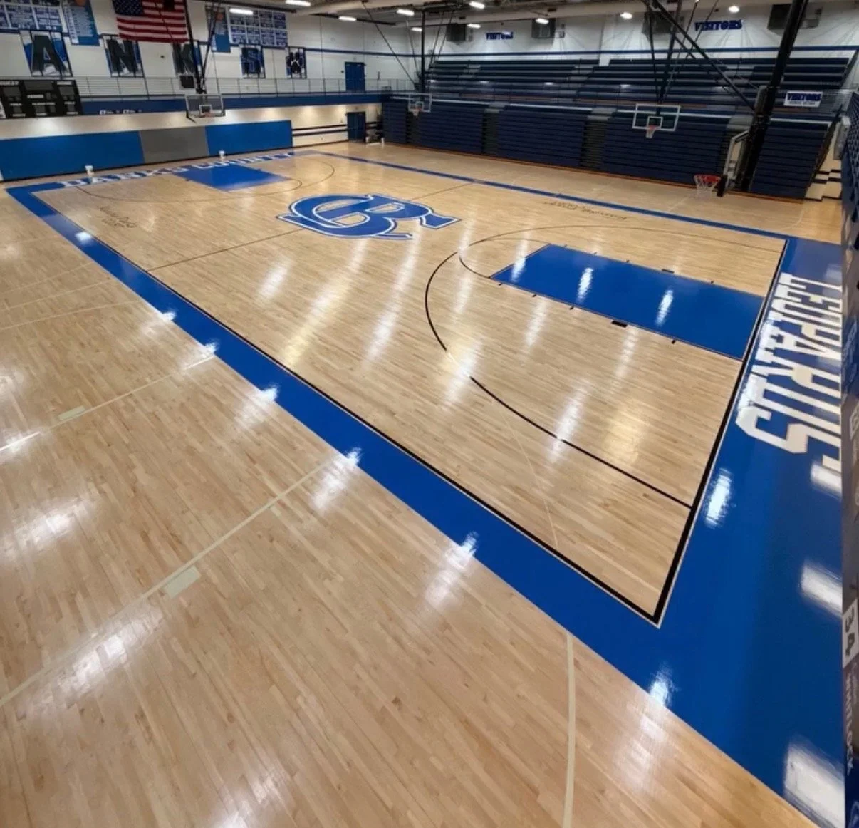 Empty basketball court with wooden flooring, blue and white markings, a large blue logo at the center, and bleachers on the sides.