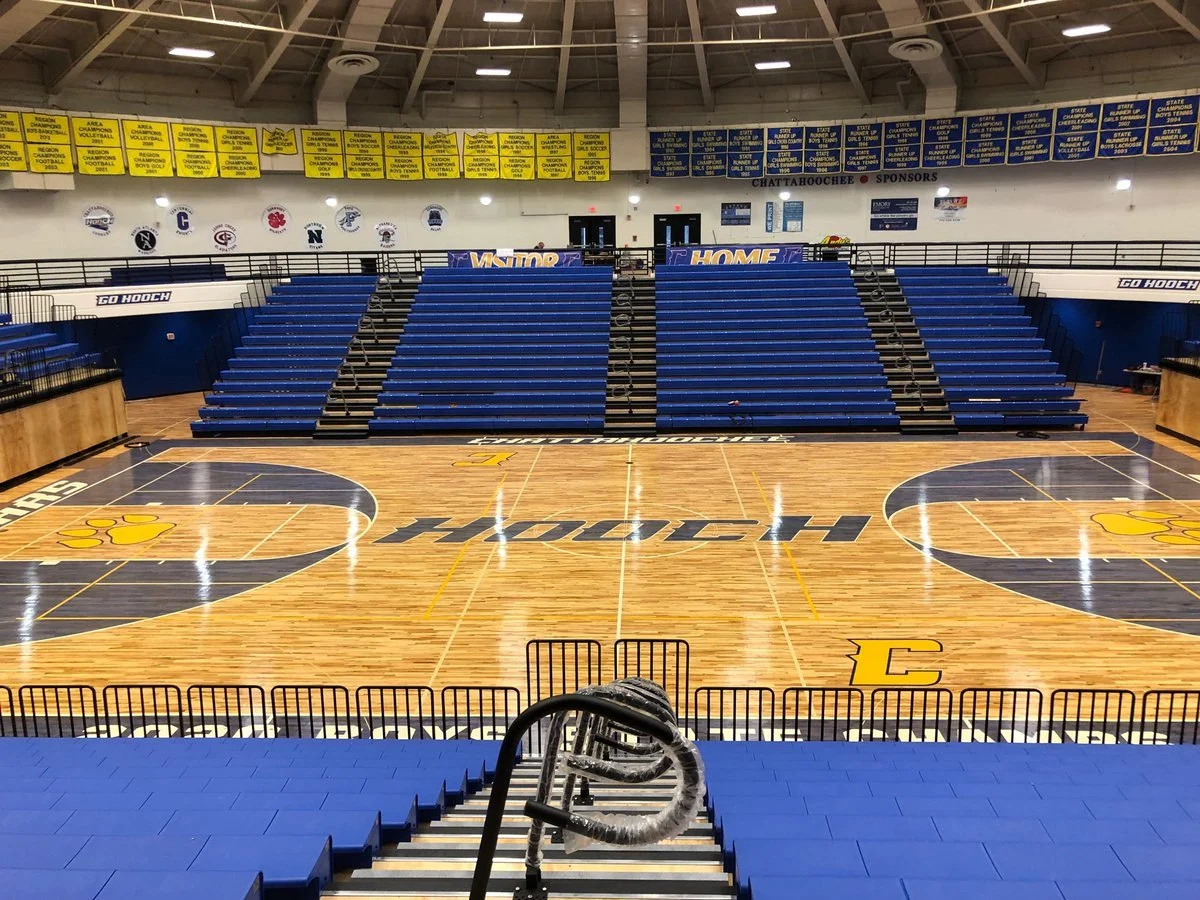 Empty basketball court with blue bleachers and banners in a gymnasium. The court has a yellow paw print logo and the word 'Hooch' in the center.