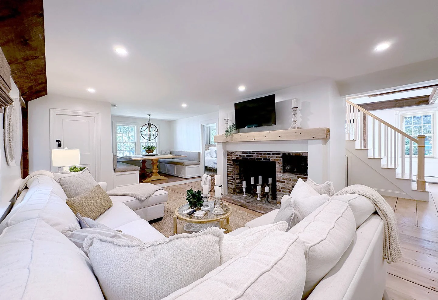 Bright living room with white sofas, a brick fireplace, mounted flat-screen TV, staircase, and a cozy dining nook.