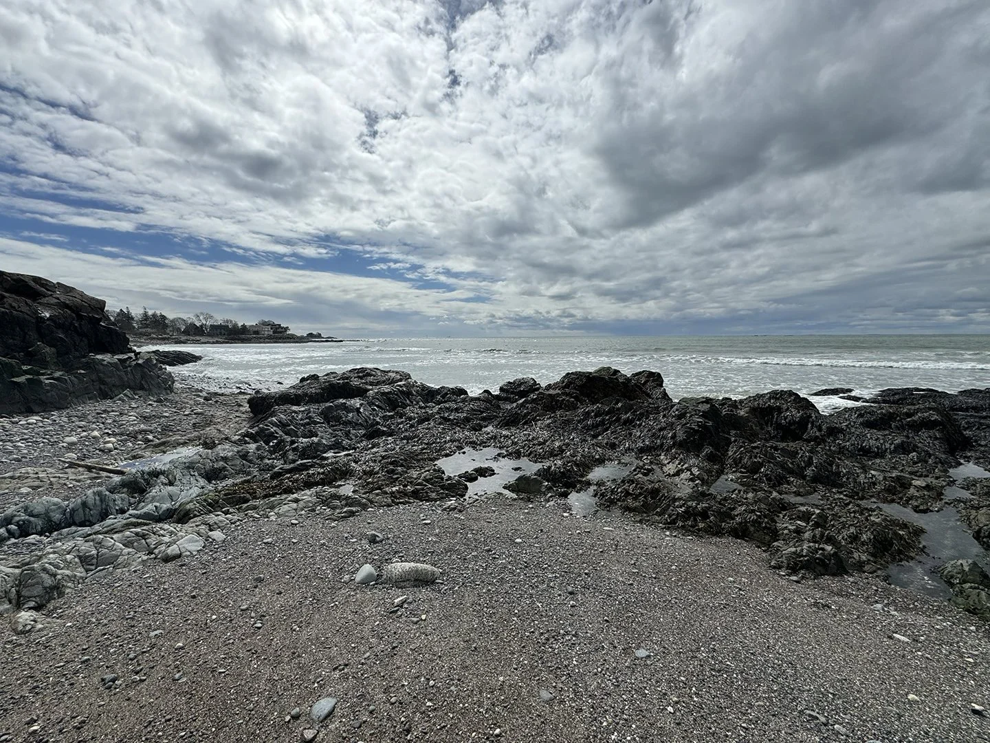 A rocky beach scene with dark rocks in the foreground, a cloudy sky overhead, and ocean waves in the background. There are some buildings visible on the distant shoreline.