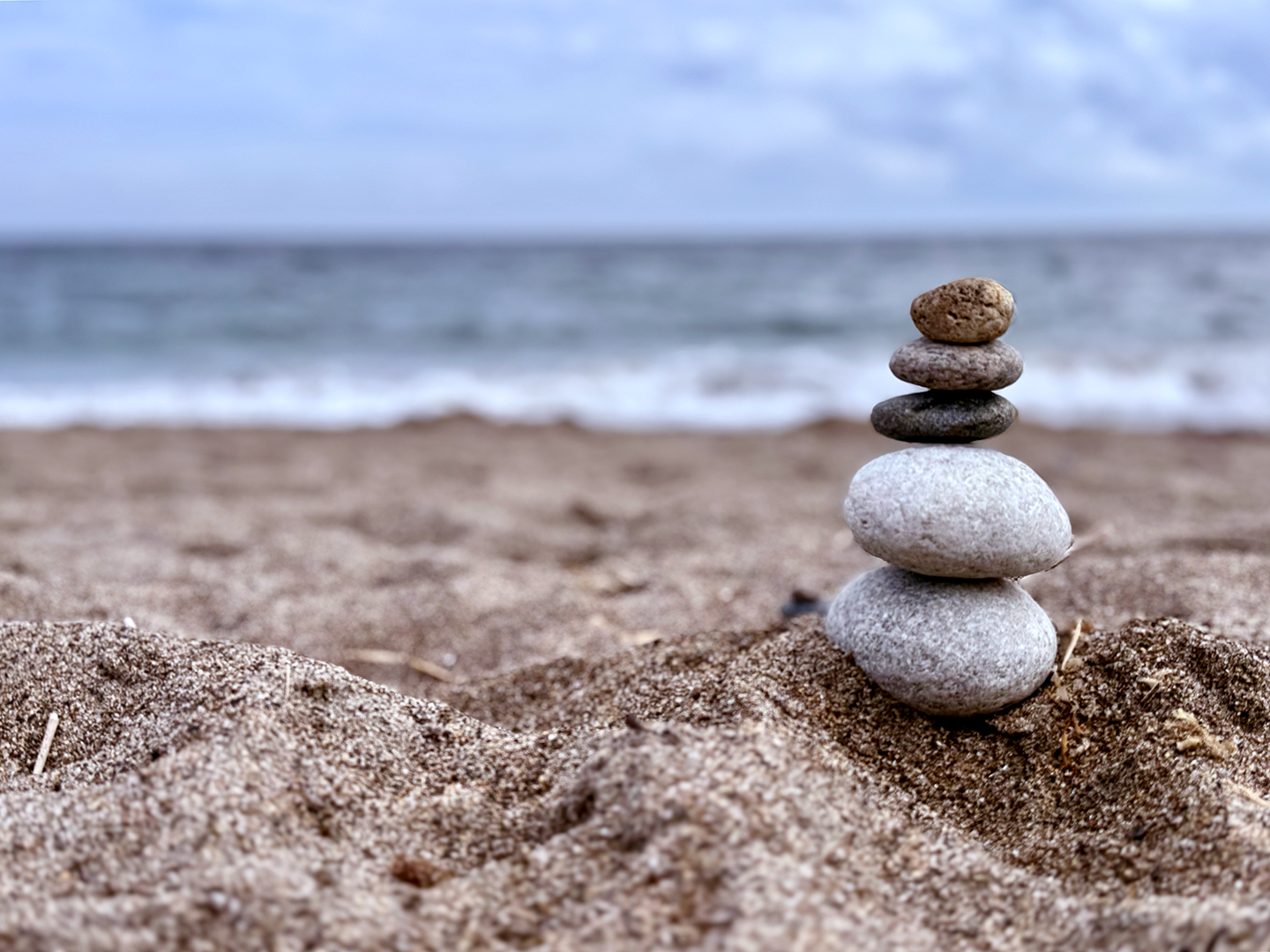 Stacked stones on sandy beach near ocean with cloudy sky in the background.