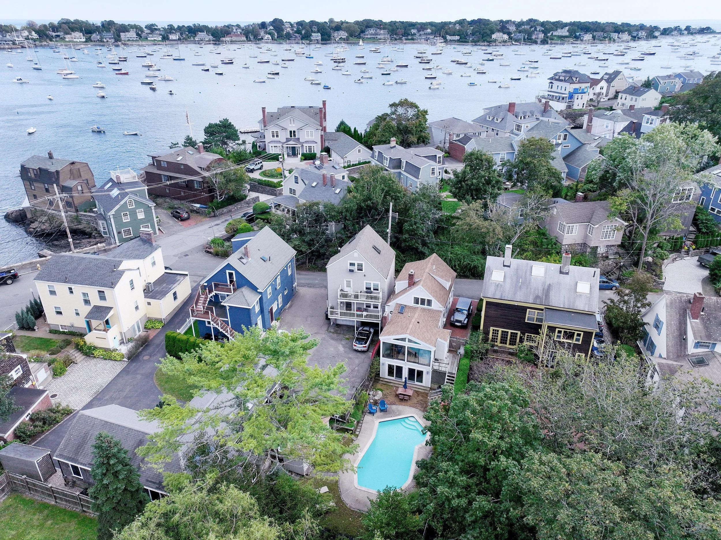 Aerial view of a coastal neighborhood with houses, a swimming pool, trees, and a marina filled with boats.