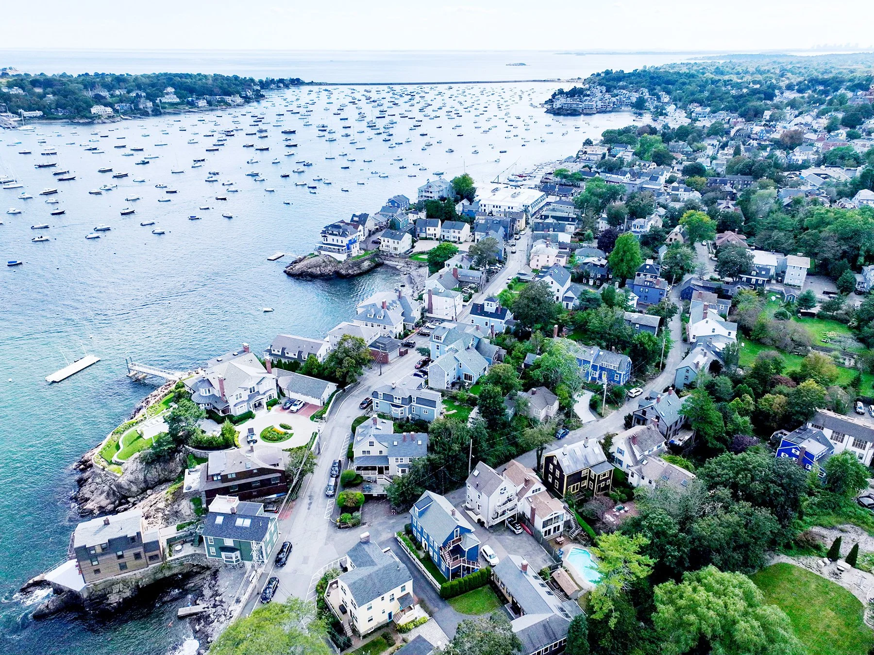 Aerial view of a coastal residential neighborhood with multiple houses, green trees, and boats anchored in the water.