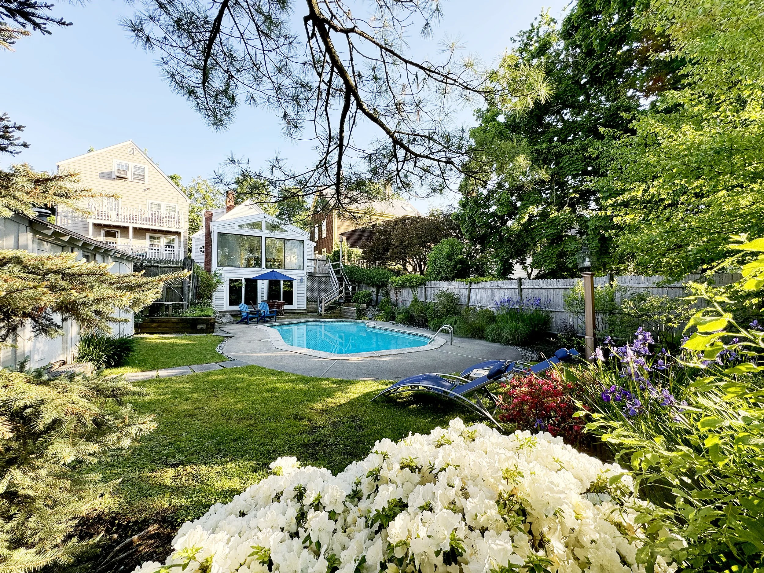 A backyard with a swimming pool, surrounded by green grass, trees, and colorful flowers. There are two lounge chairs and a small table near the pool, with a house and sunroom in the background.