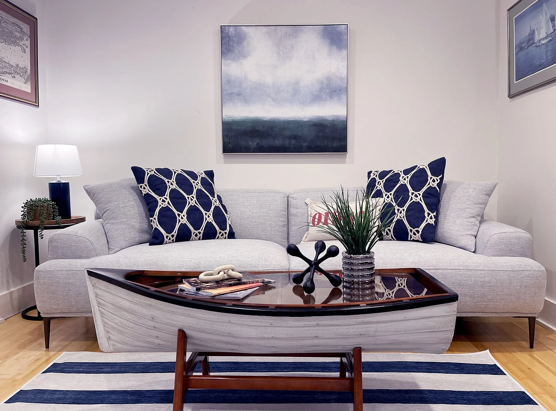 A living room with a light gray sofa decorated with navy and patterned throw pillows, a wooden coffee table with a decorative sculpture and books, a side table with a lamp and plant, and a striped rug on hardwood floor. Artwork hangs on white walls.
