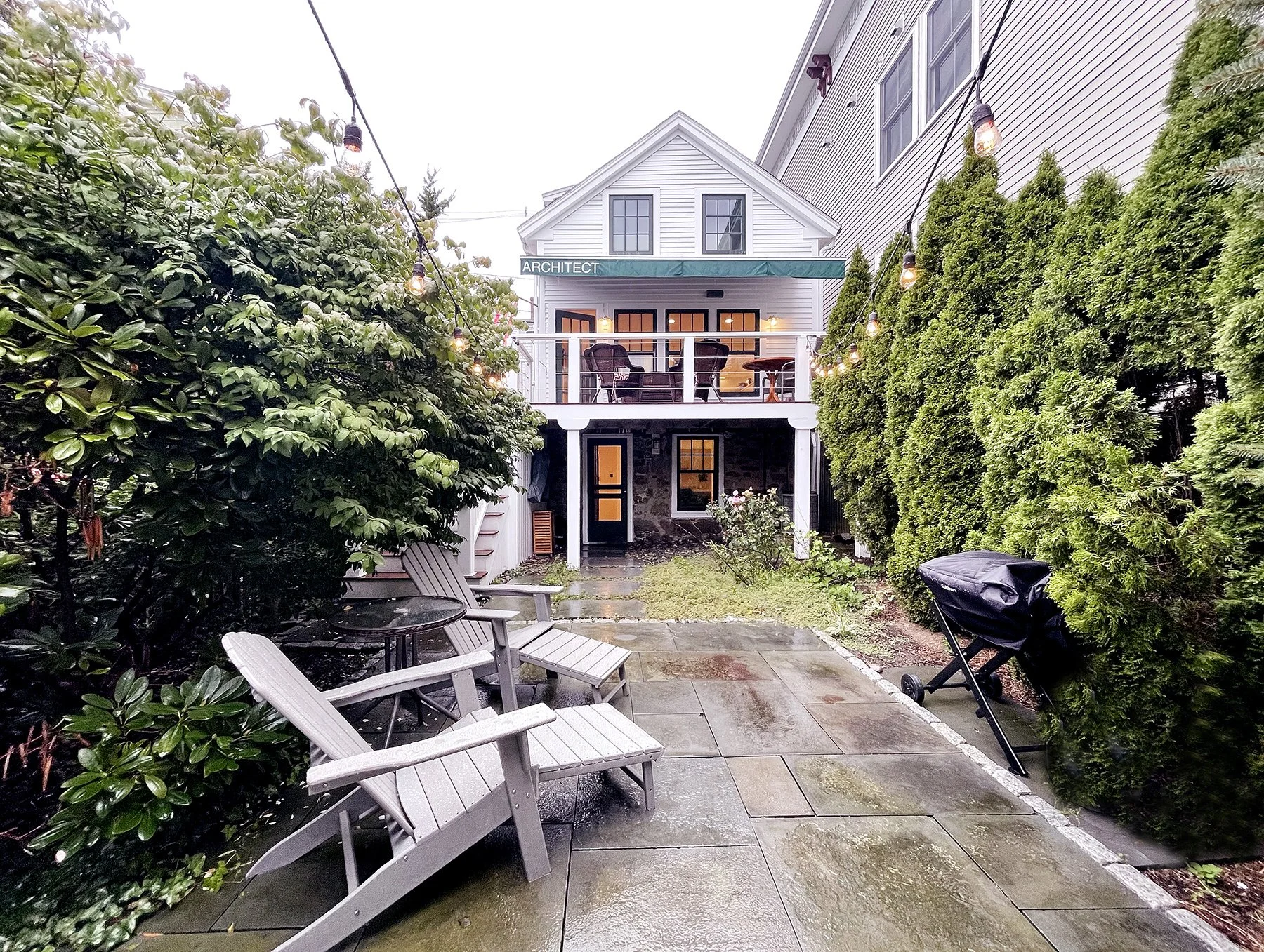 Backyard patio with wooden benches and Adirondack chairs, a barbecue grill, and string lights, with a two-story house featuring a small balcony and green hedges on either side.