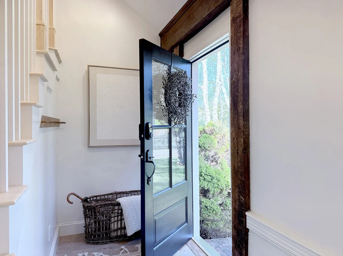 Open front door of house revealing a view outside with trees and bushes, white interior walls, a framed white textured artwork, a woven basket with a white blanket, a staircase on the left, and a focus on the doorway with a wreath hanging on the door