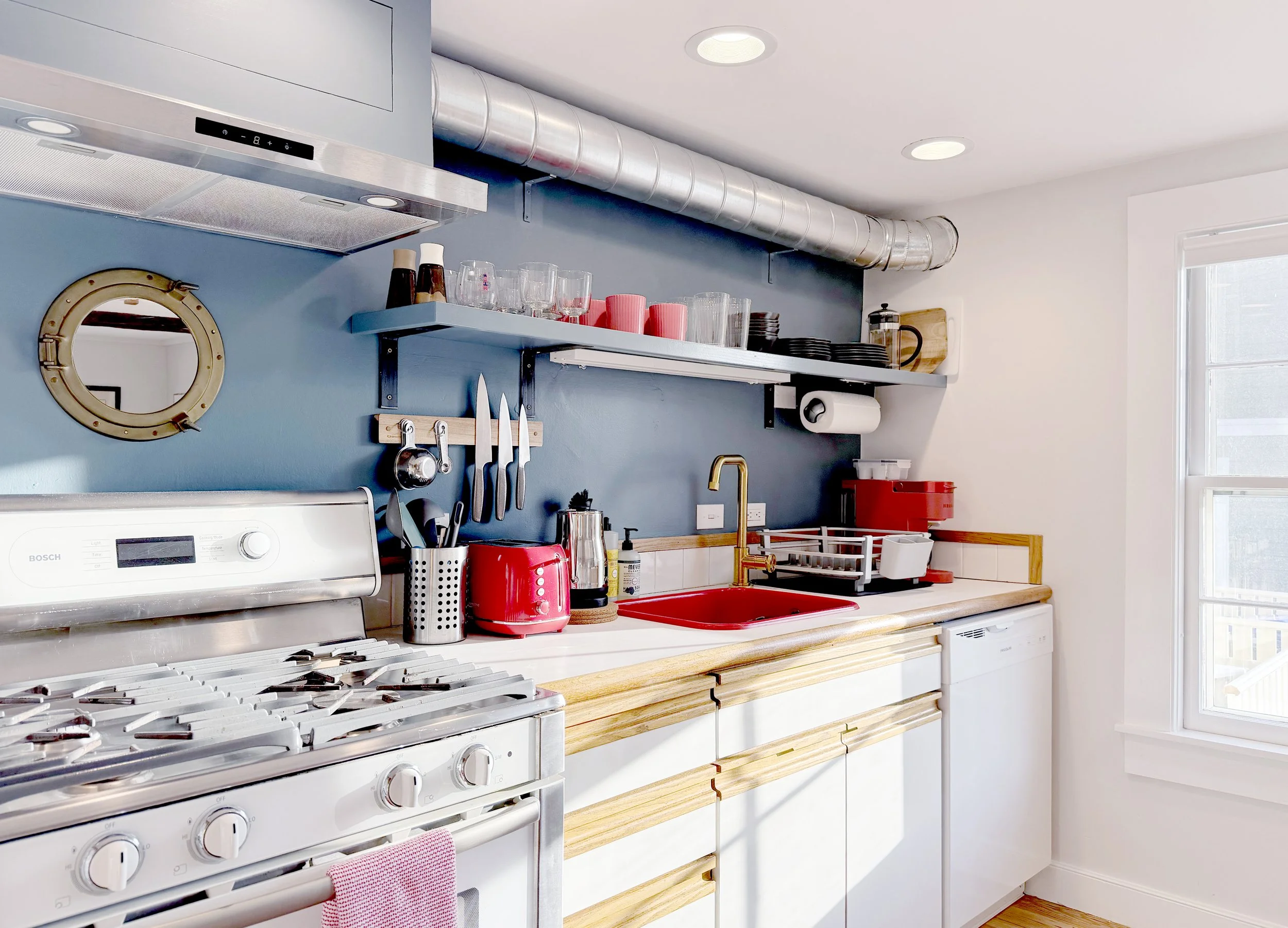 Modern kitchen with a white stove, red sink, and blue accent wall with shelves holding glasses, cups, and plates. Kitchen tools and appliances are on the countertop, with a window on the right providing natural light.