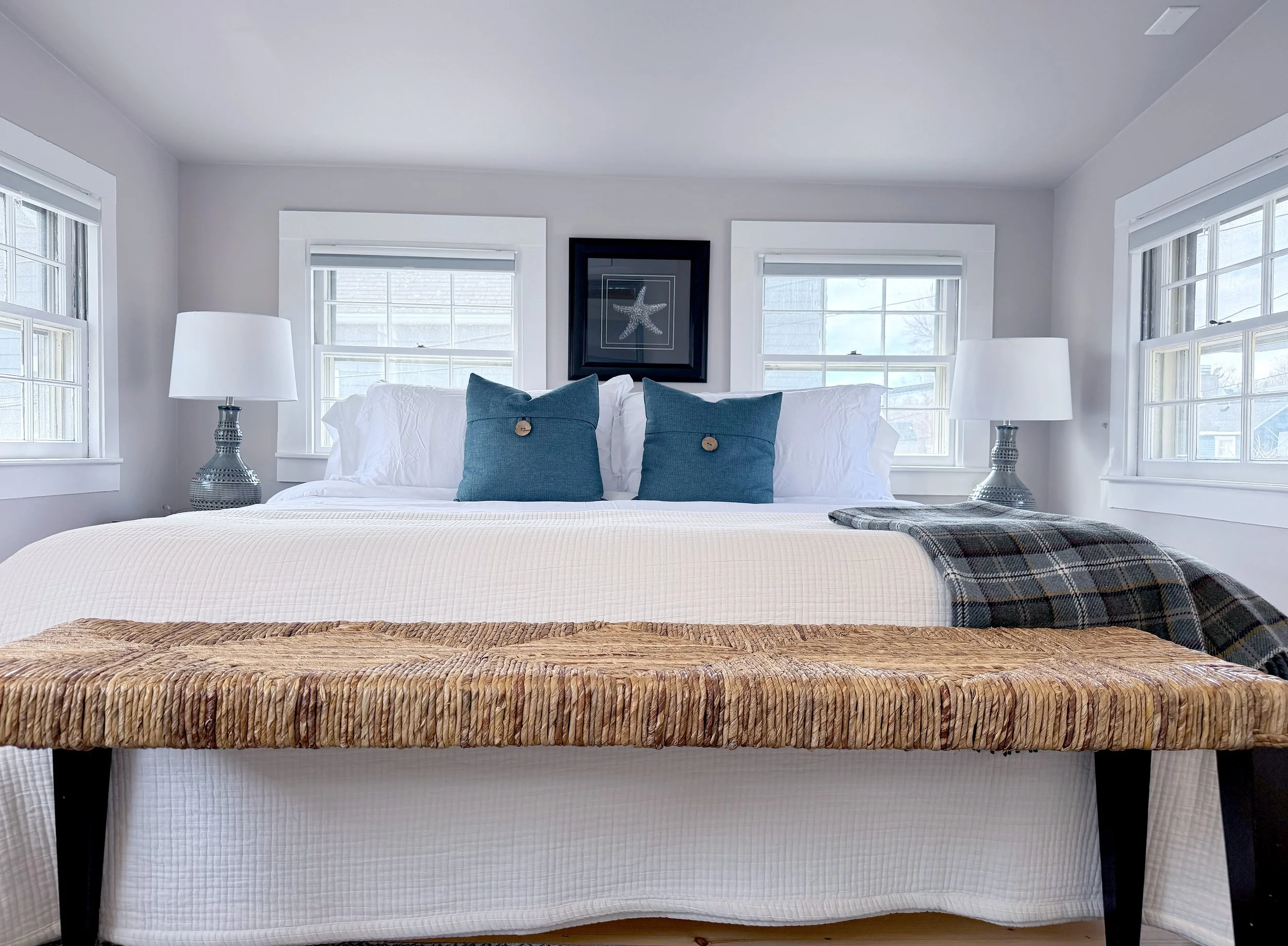 A neatly made bed with white bedding, two blue pillows with buttons, two white pillows, and a plaid blanket, flanked by two matching lamps, in a bright room with three windows and a framed artwork of a starfish.