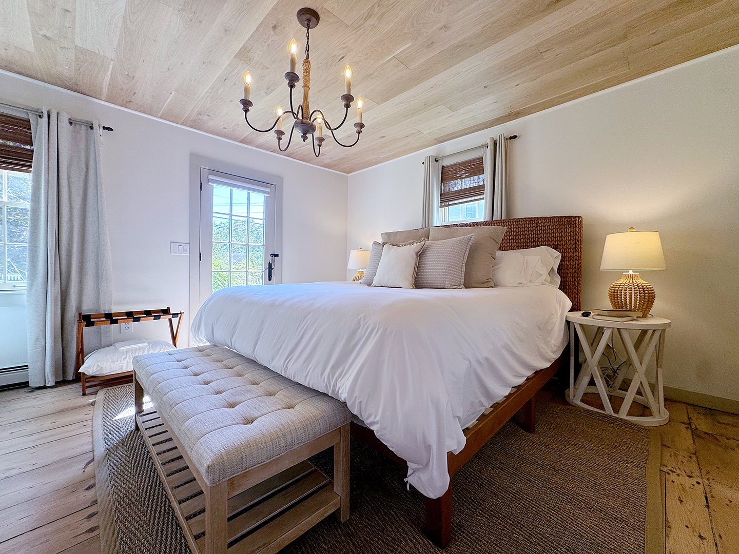 Bedroom with a large bed with white linens, multiple pillows, wooden headboard, side table with lamp, and a bench at the foot of the bed. There are windows with curtains, a door with a window, and a chandelier on the ceiling.