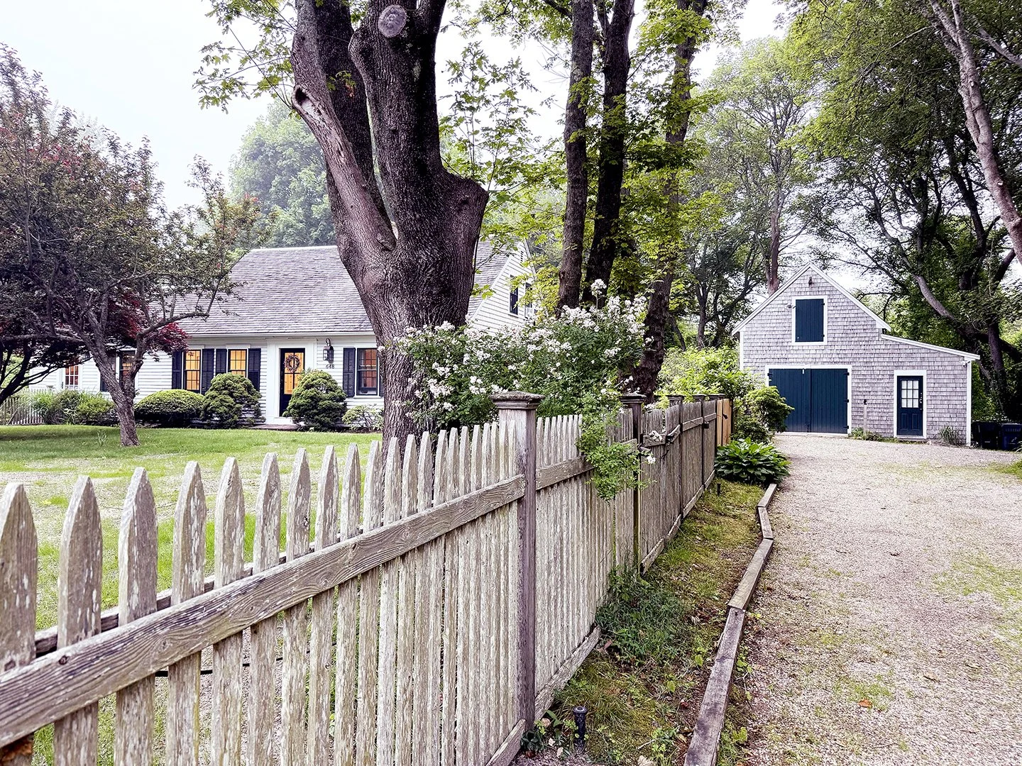 A white house with black shutters and a manicured lawn, next to a wooden fence, large trees, and a gravel driveway leading to a gray shed with blue doors.