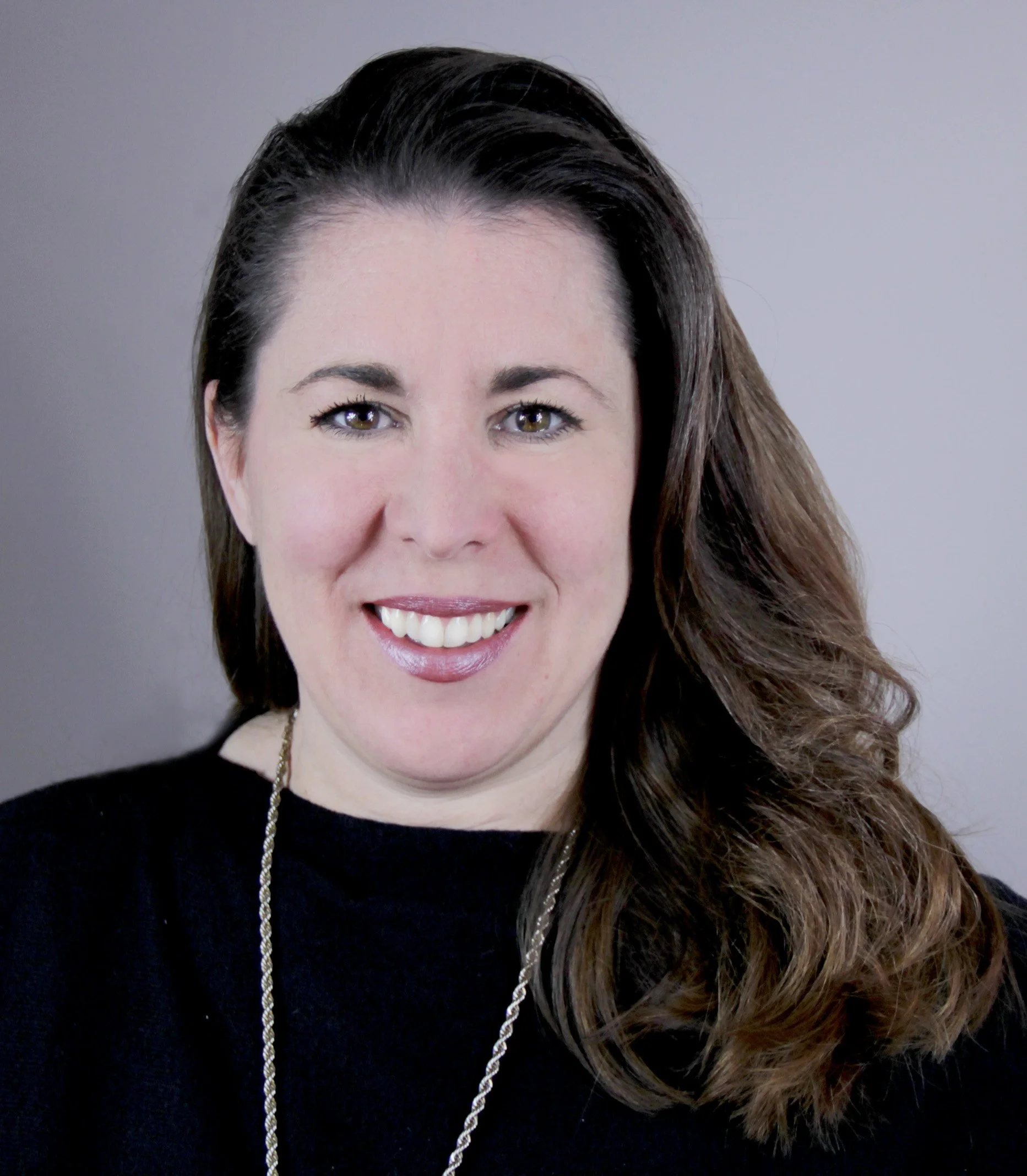 A woman with long brown hair smiling at the camera, wearing a black top and a necklace, against a neutral background.