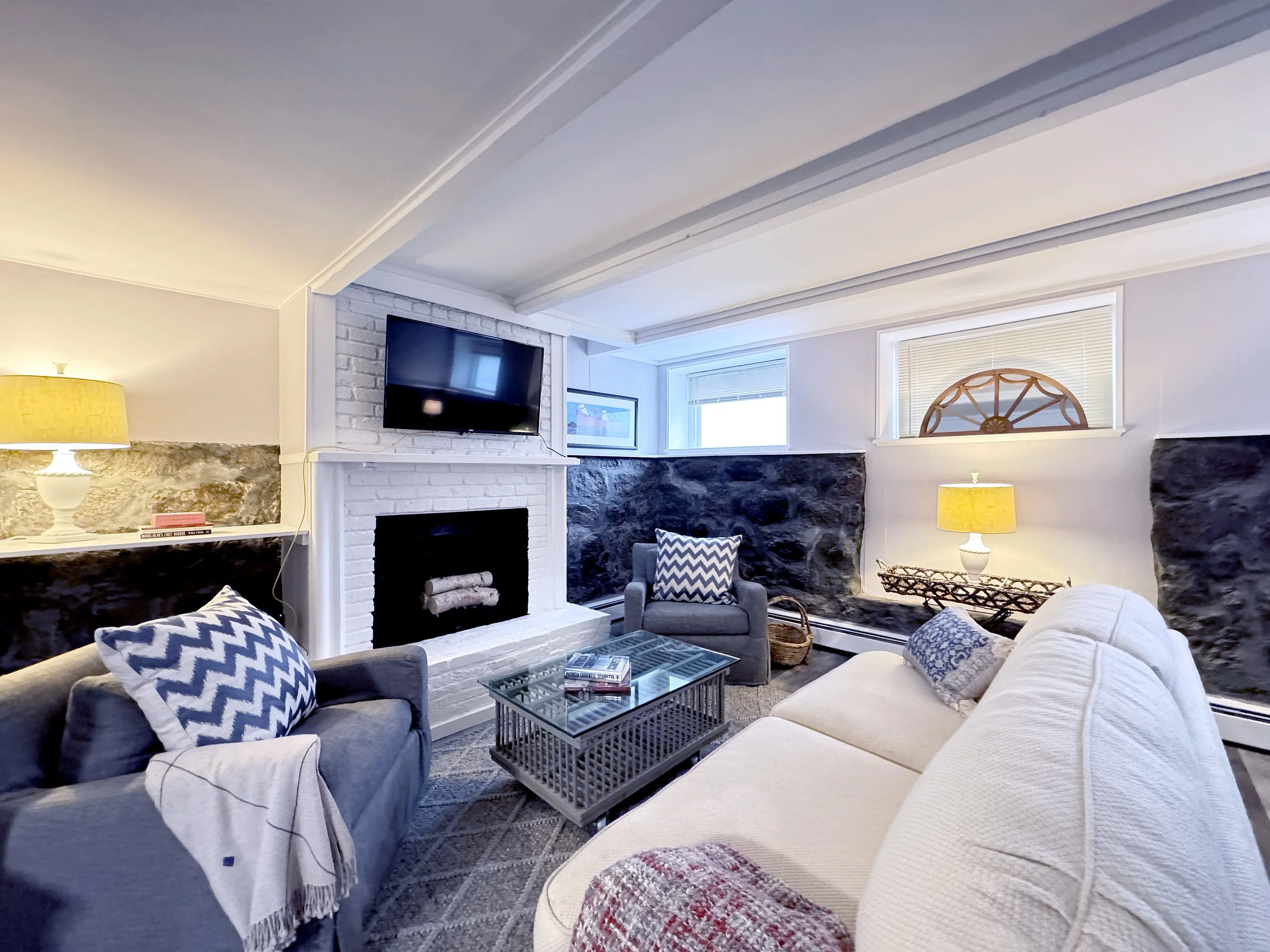 Cozy living room with white brick fireplace, mounted flat-screen TV, cushioned armchair, sofa with patterned pillows, glass coffee table, textured walls, and yellow lamps.